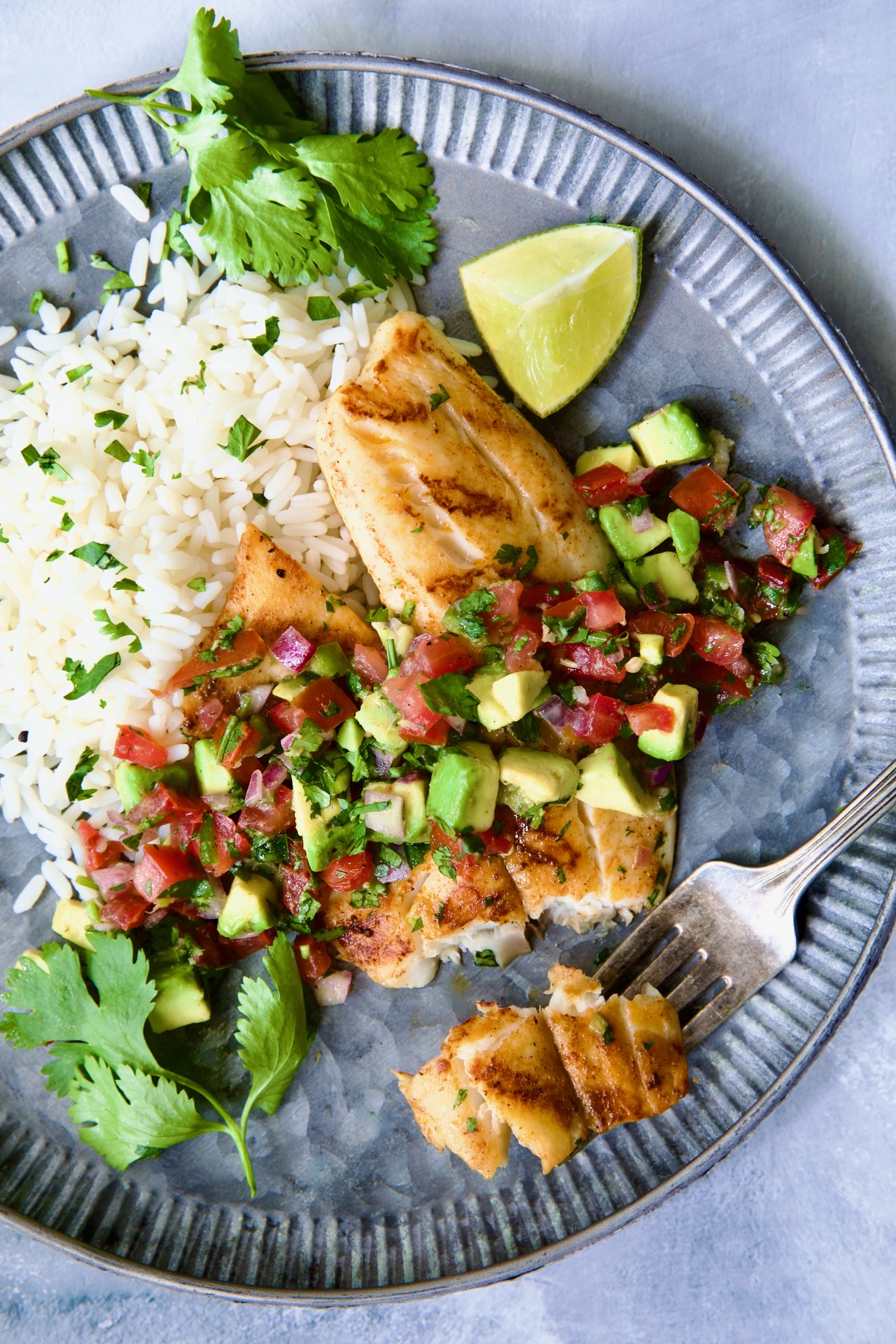 Grilled Tilapia with Tomato - Avocado Salsa on round metal plate with fork having cut some off ready to eat.