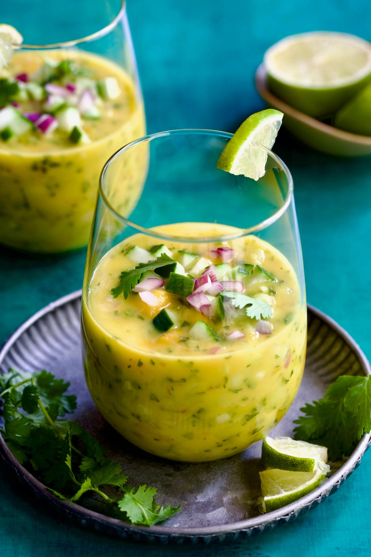 Two glasses of Mango Gazpacho with one on metal tray garnished with cilantro and lime wedges.