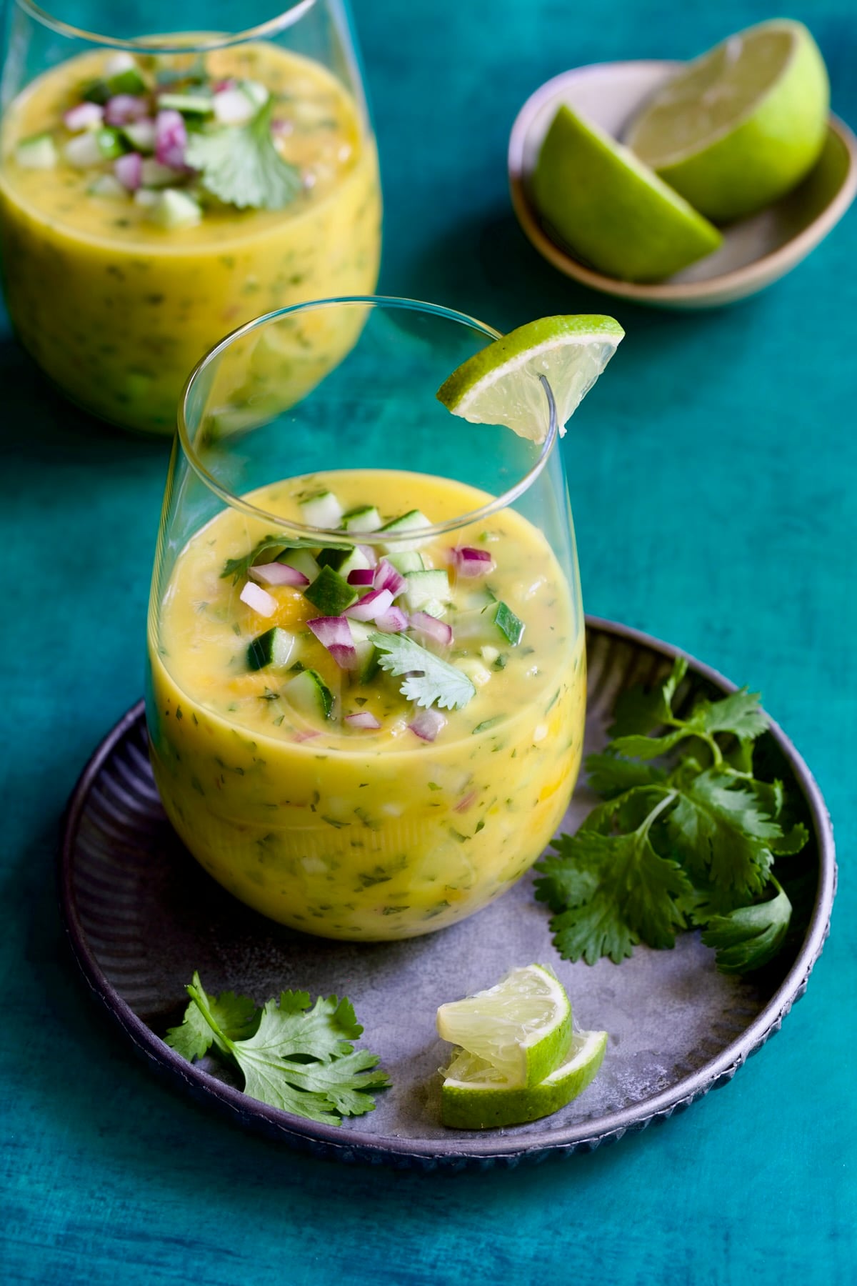 Two glasses of Mango Gazpacho with one on metal tray garnished with cilantro and lime wedges.