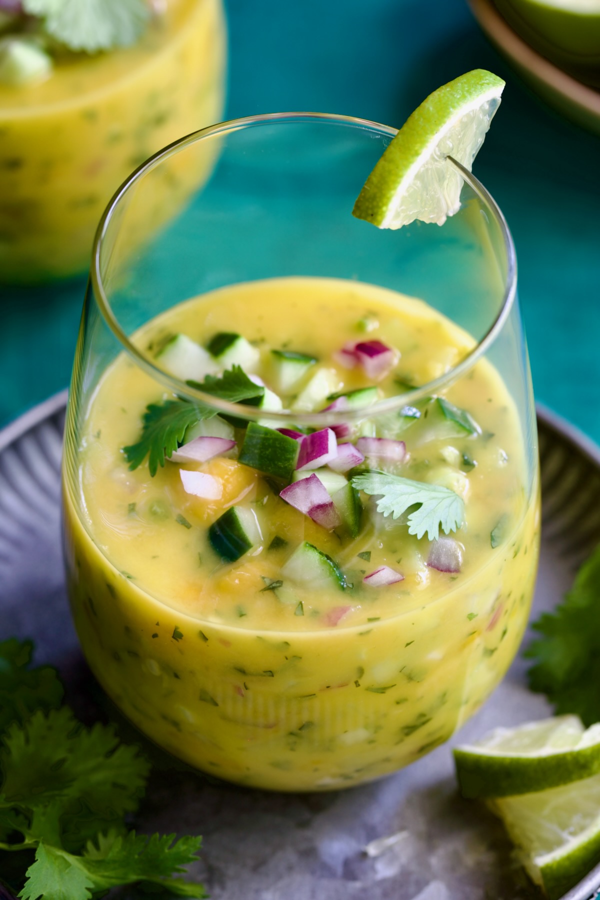 One glass of Mango Gazpacho with one on metal tray garnished with cilantro and lime wedges.