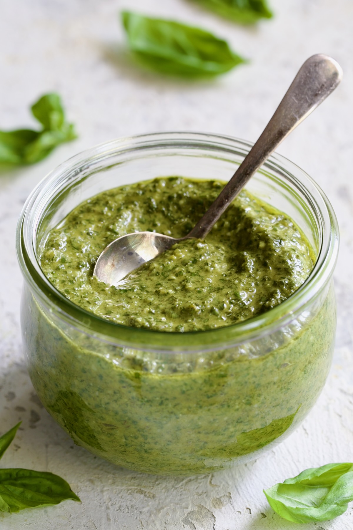 Jar of Pesto Genovese with spoon with fresh basil leaves scattered about.