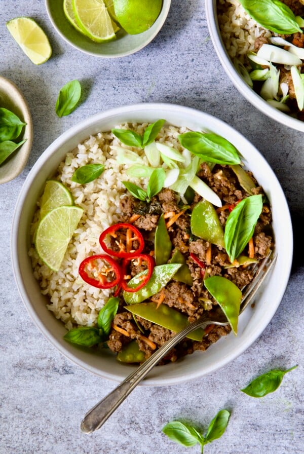 Thai Basil Beef in white bowl garnished with lime wedges, fresh basil and Fresno chiles.