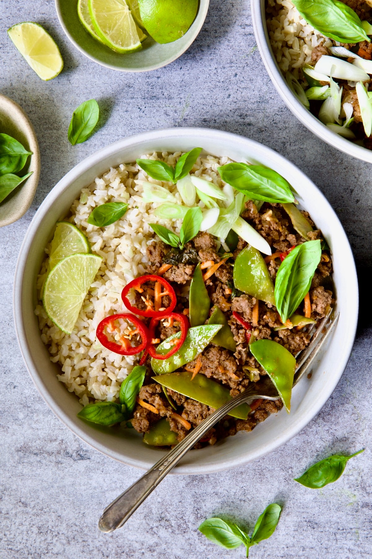 Thai Basil Beef in white bowl garnished with lime wedges, fresh basil and Fresno chiles.
