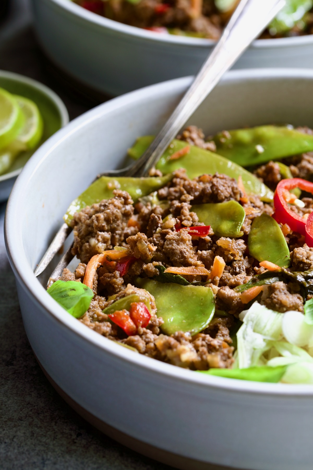 Thai Basil Beef in white bowl garnished with lime wedges, fresh basil and Fresno chiles with fork.
