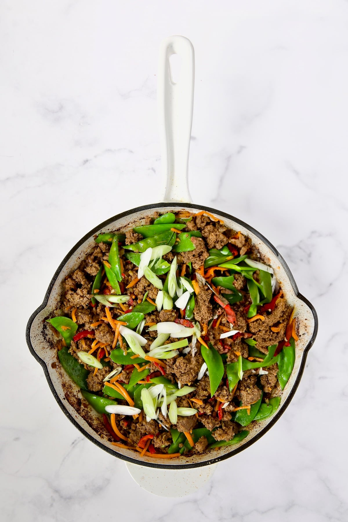 Cooked ground beef, Fresno chiles, shredded carrots, snow peas and scallions in white enamel cast iron skillet.