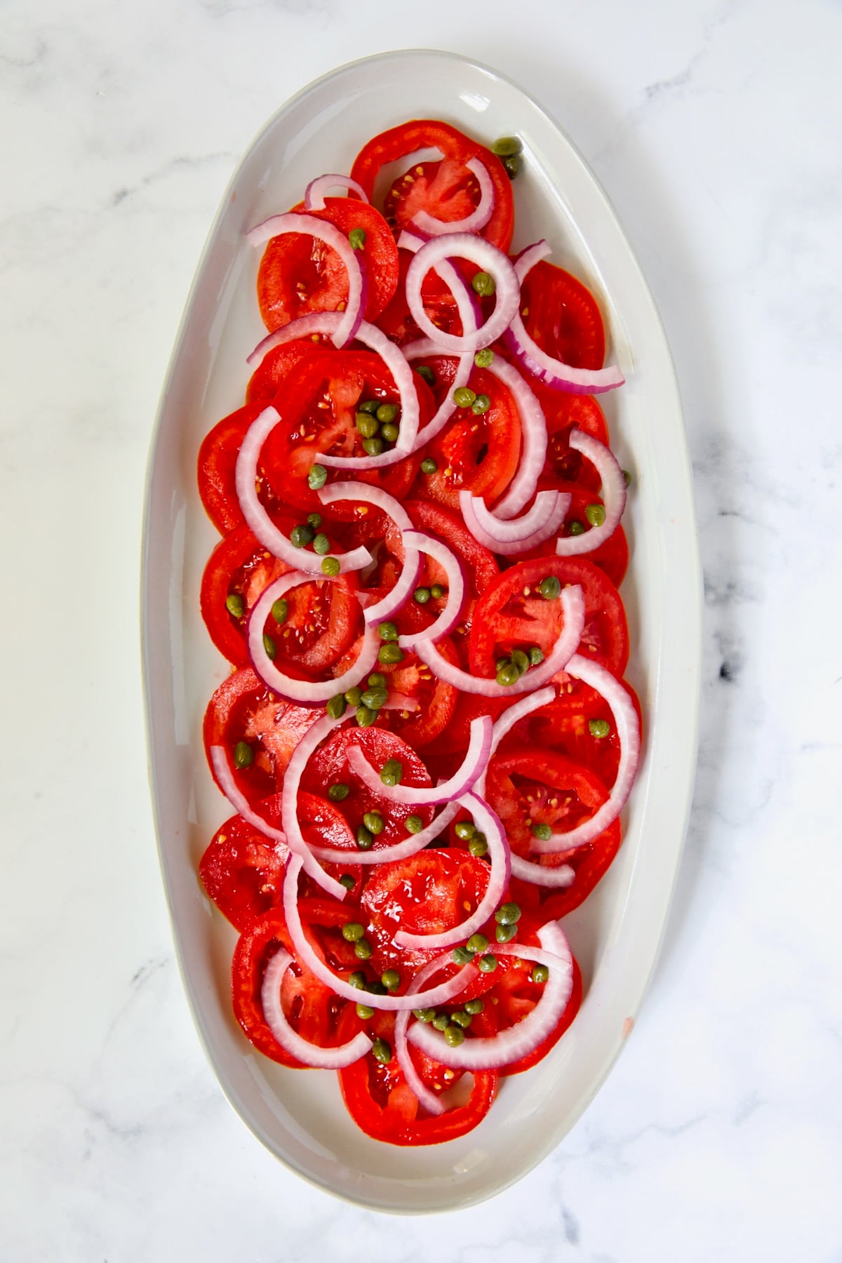 Sliced tomatoes, red onion slices and capers on white oval platter.