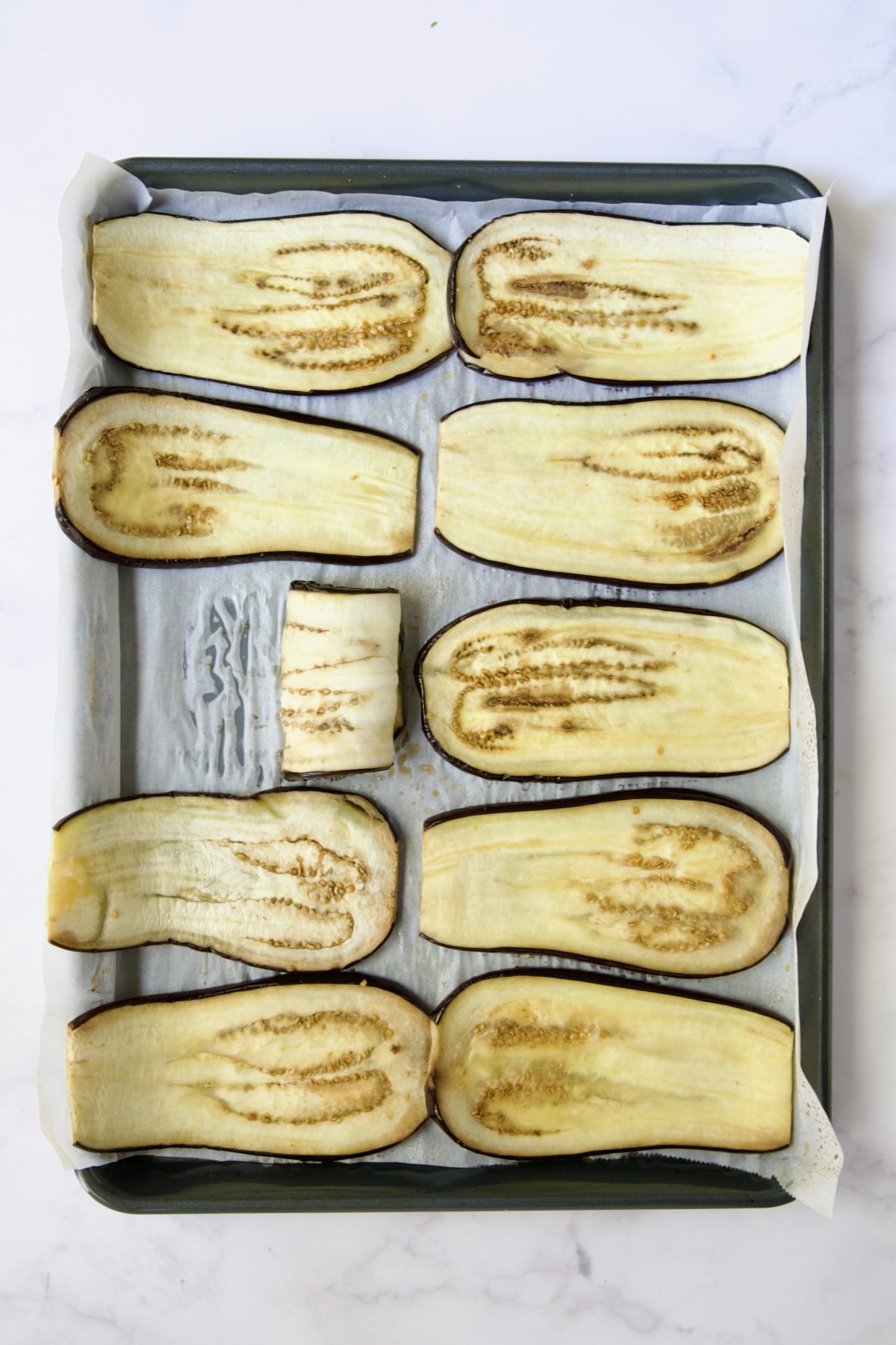 Ten baked eggplant slices on parchment paper-lined baking sheet with one rolled up to show pliability.