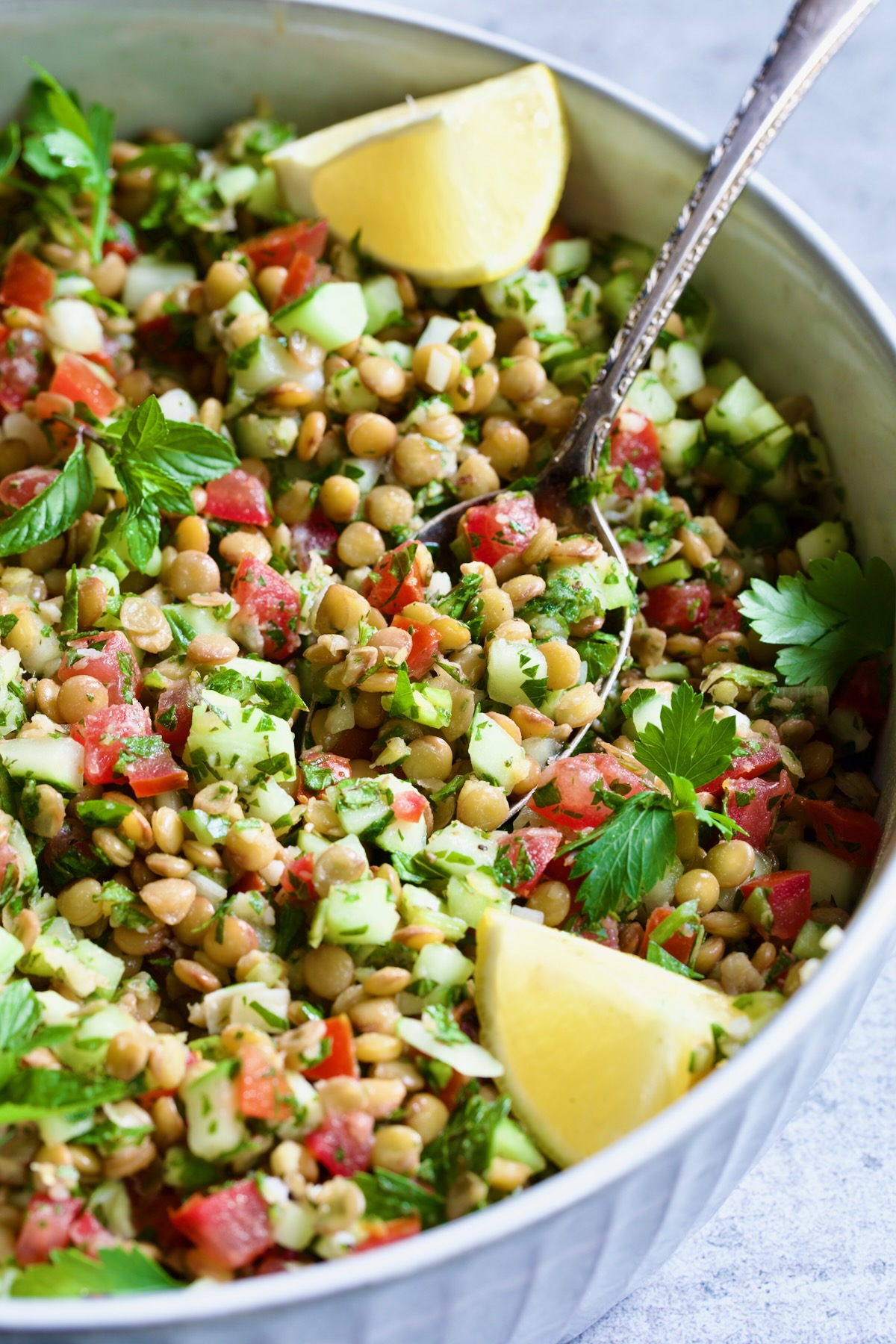 Lentil Tabbouleh in white bowl with serving spoon garnished with fresh mint sprigs and lemon wedges.