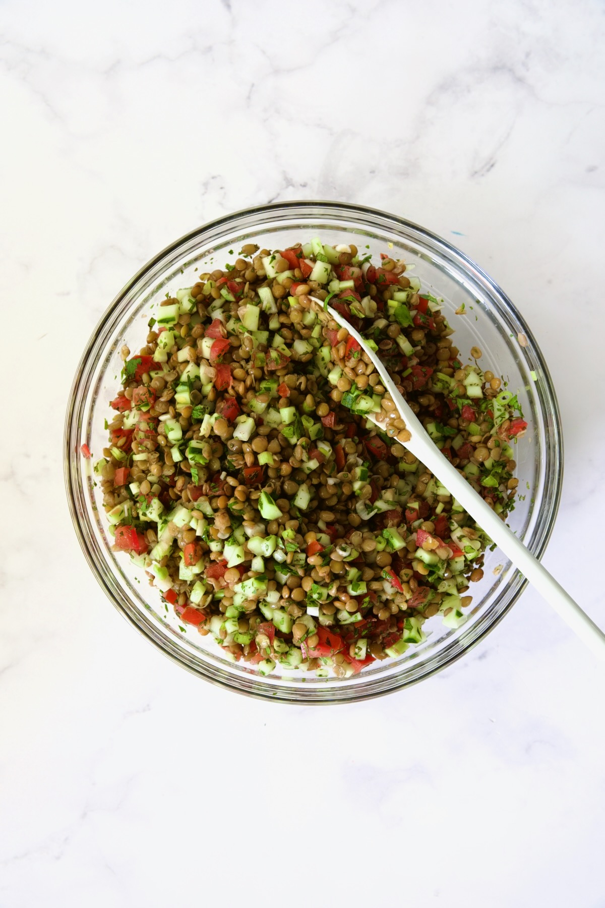 Ingredients for Lentil Tabbouleh in glass bowl after being mixed together with white mixing spoon.
