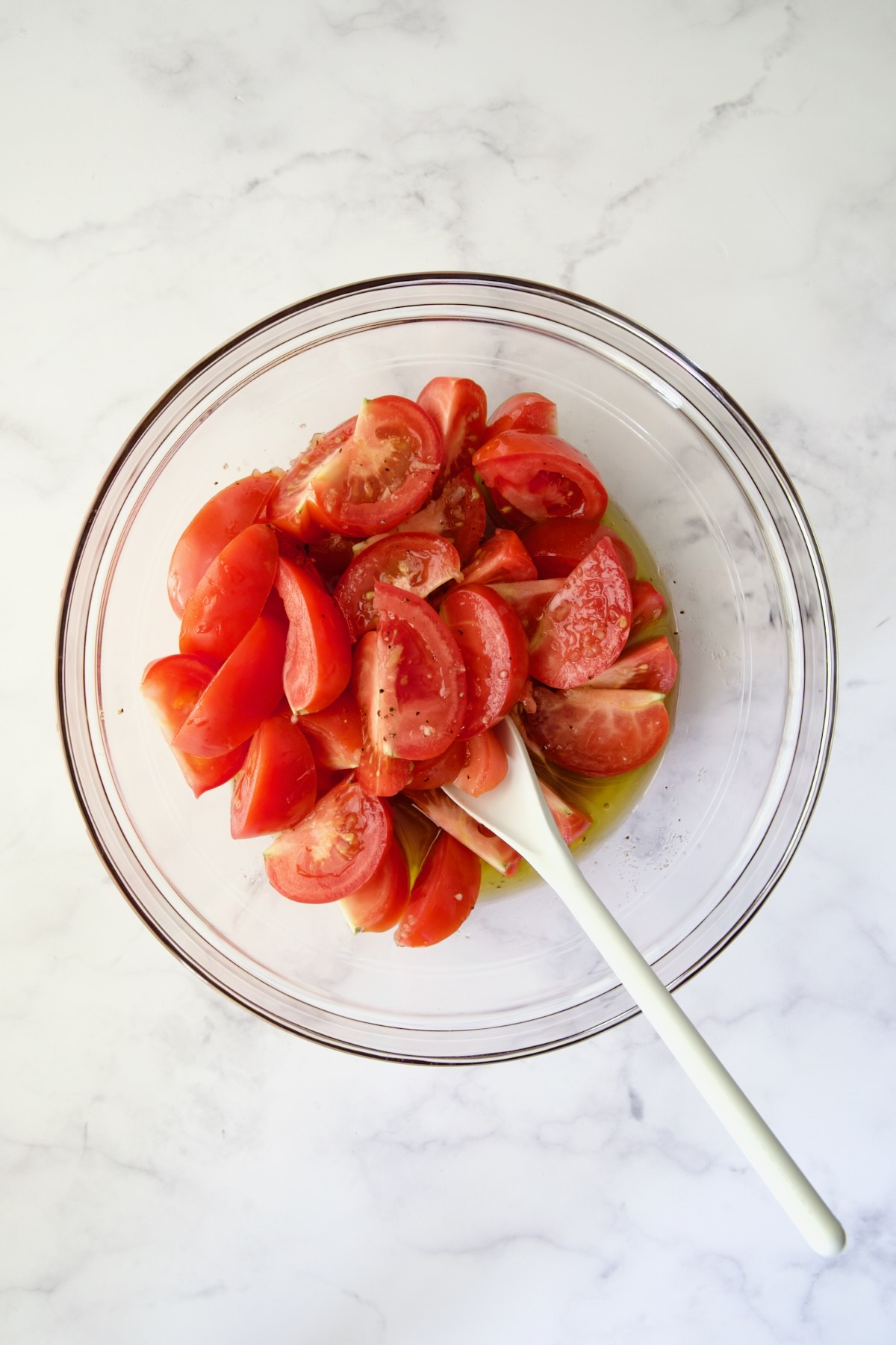 Tomato wedges in glass bowl being stirred with white mixing spoon.