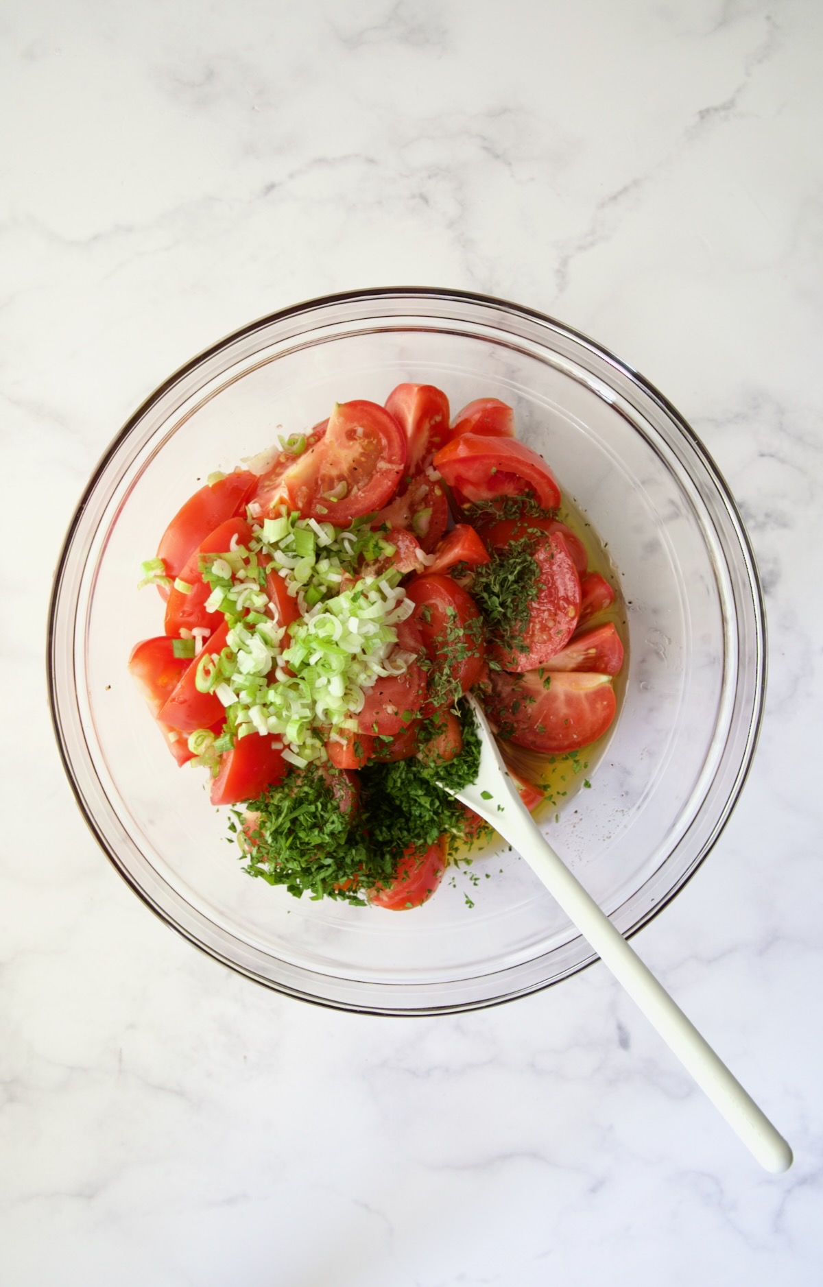 Herbs and scallions added to tomato wedges in glass bowl.