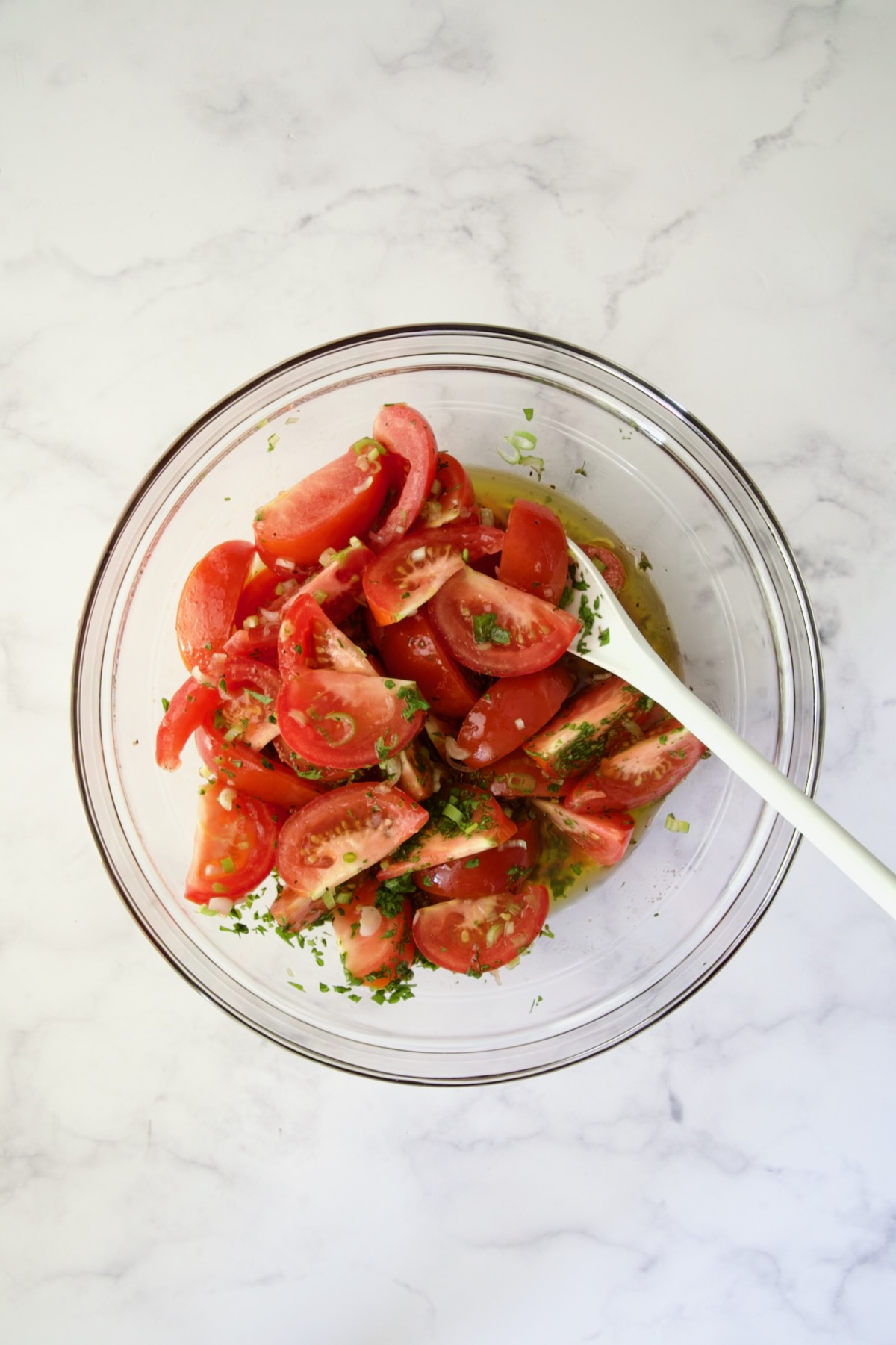 Tomato wedges with dressing, scallions and herbs being stirred in glass bowl with white mixing spoon.