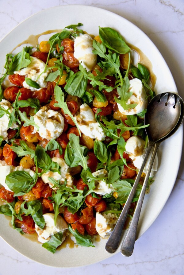 Roasted Cherry Tomato Burrata Caprese Salad on white oval platter with serving utensils.