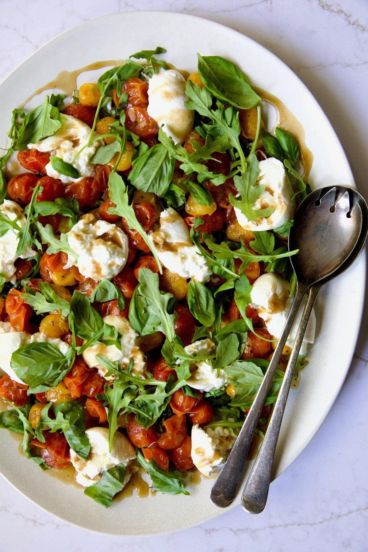 Roasted Cherry Tomato Burrata Caprese Salad on white oval platter with serving utensils.
