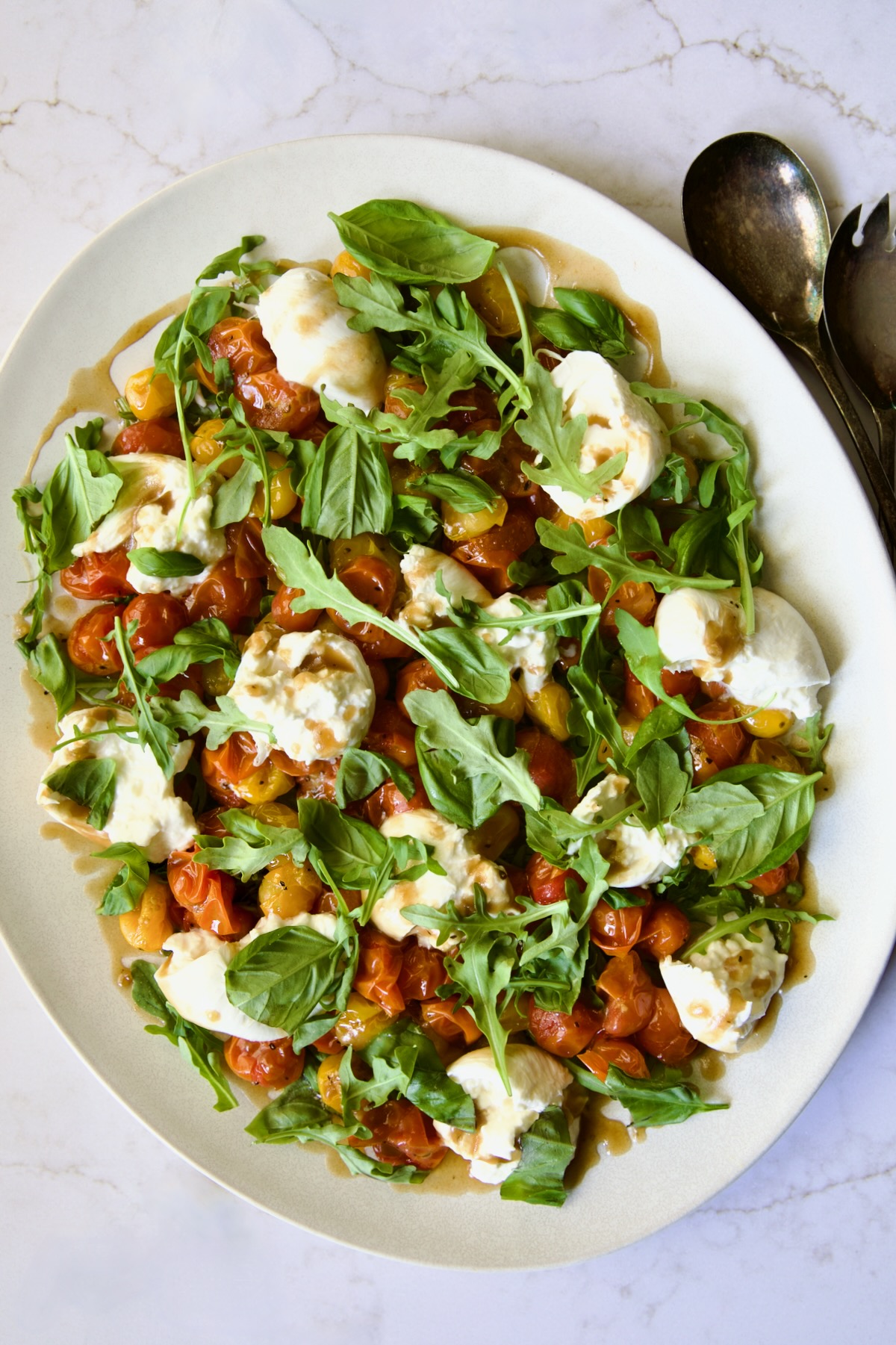 Roasted Cherry Tomato Burrata Caprese Salad on white oval platter with serving utensils.