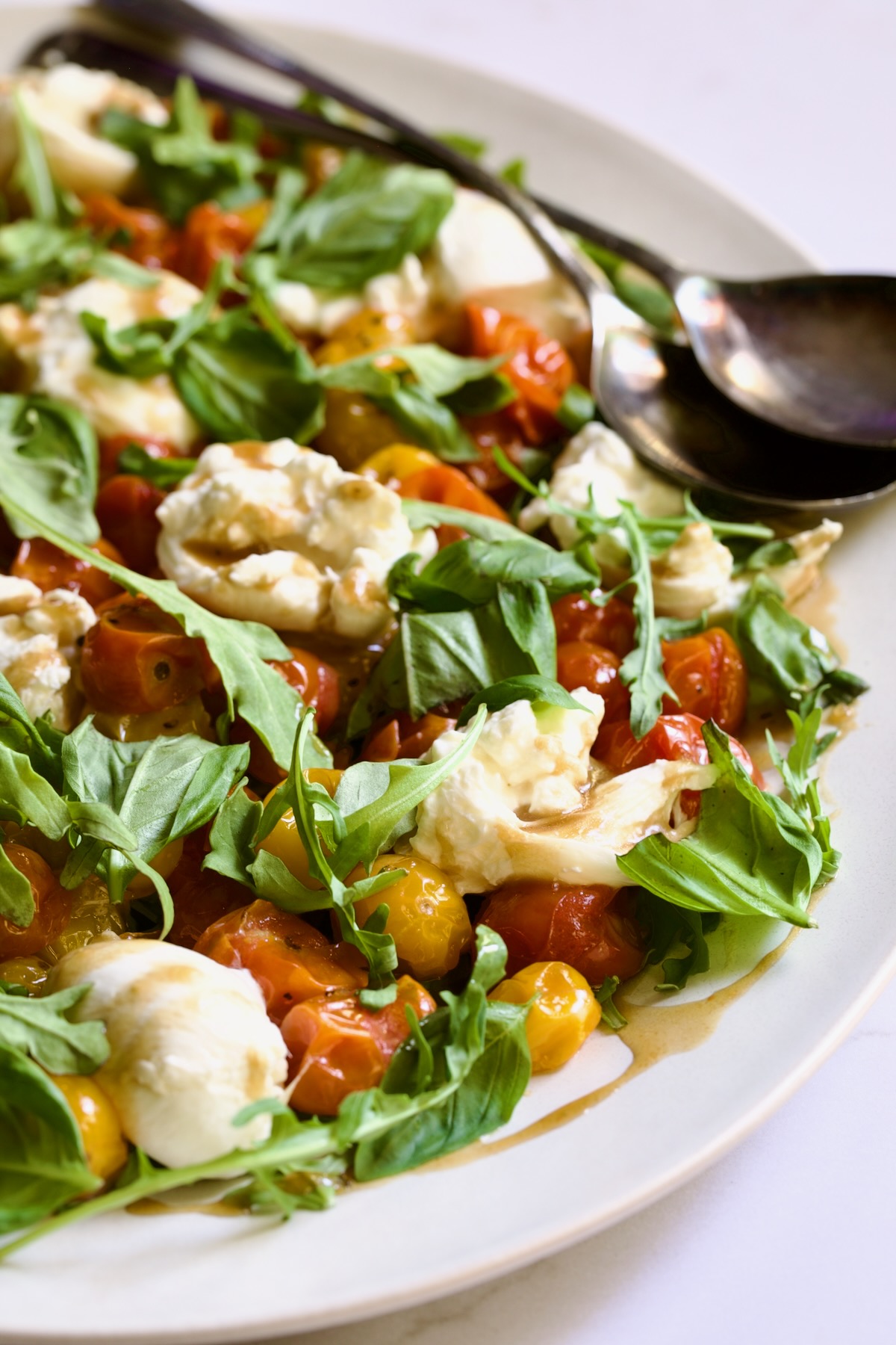 Roasted Cherry Tomato Burrata Caprese Salad on white oval platter with serving utensils.