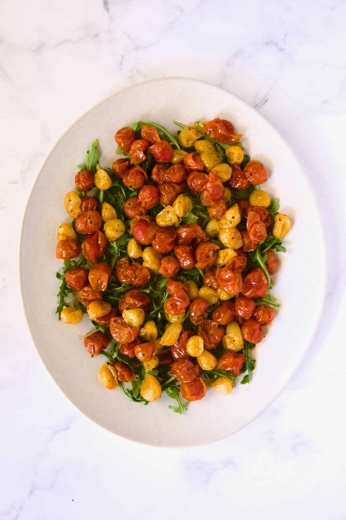 Roasted cherry tomatoes atop arugula on white oval platter.