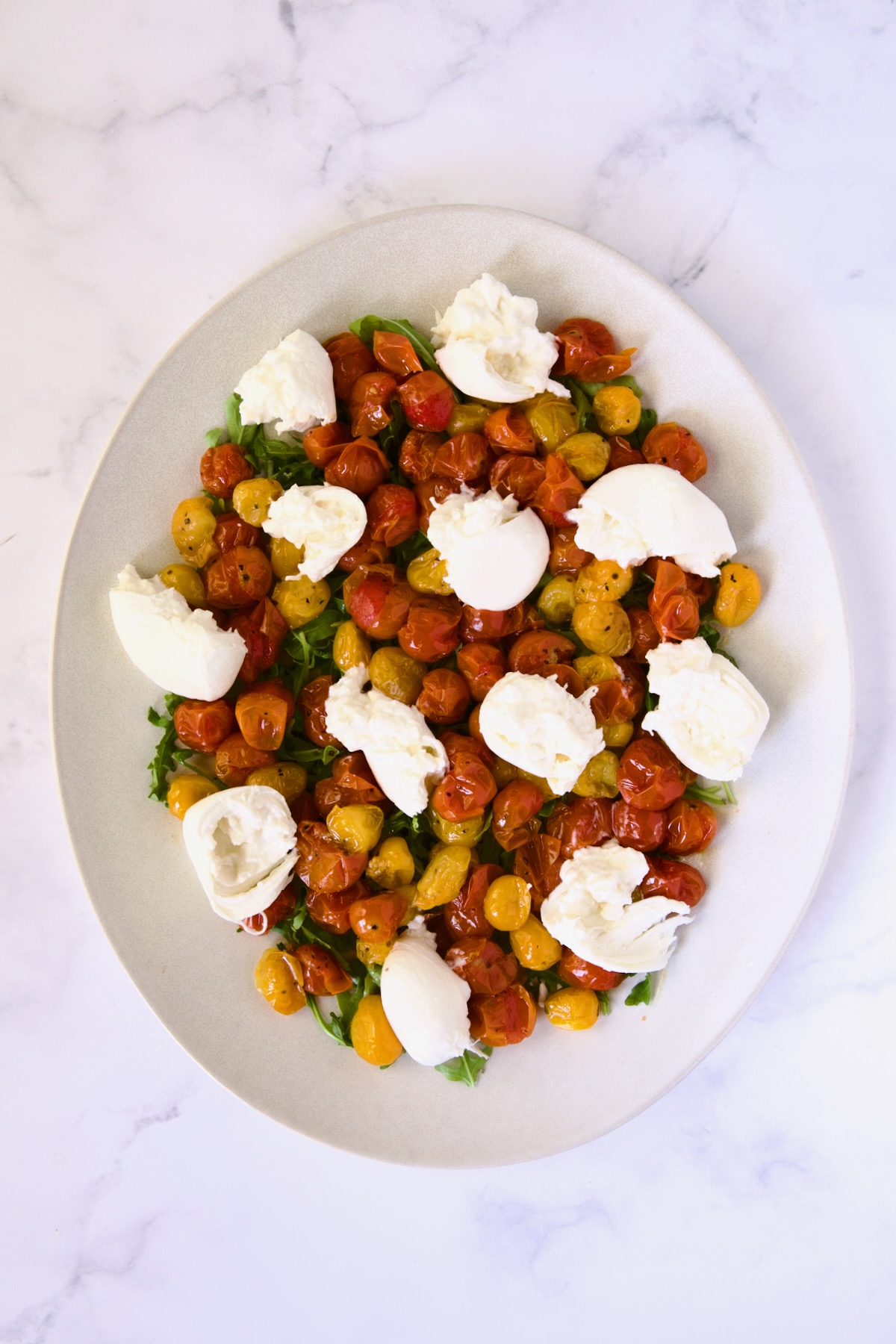 Burrata and roasted cherry tomatoes atop arugula on white oval platter.