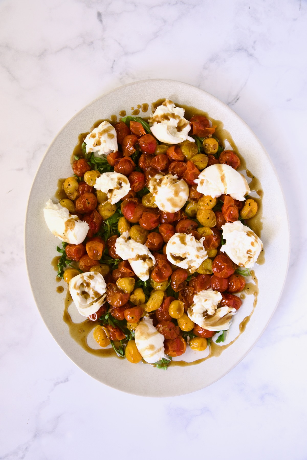 Roasted cherry tomatoes, arugula and burrata on white oval platter drizzled with balsamic vinaigrette.