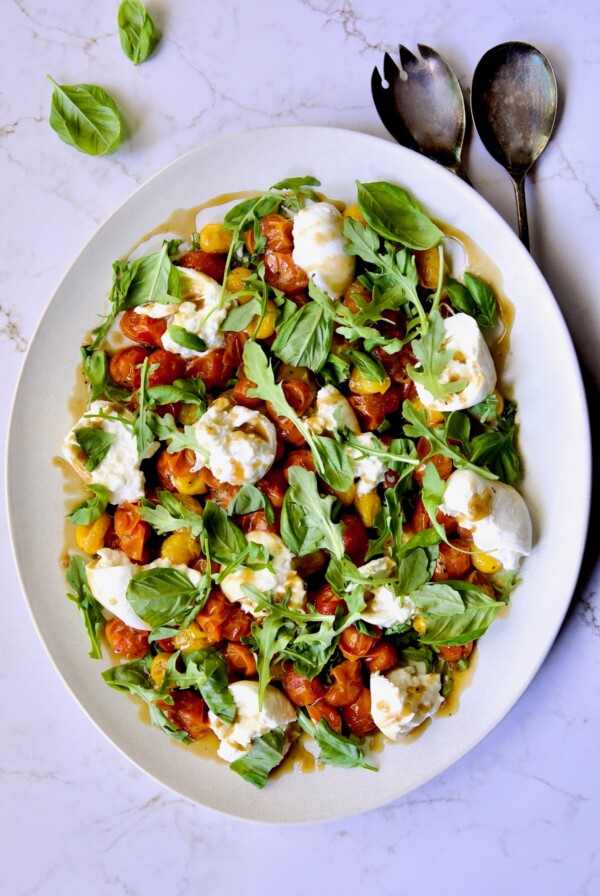 Roasted Cherry Tomato Burrata Caprese Salad on white oval platter with serving utensils.