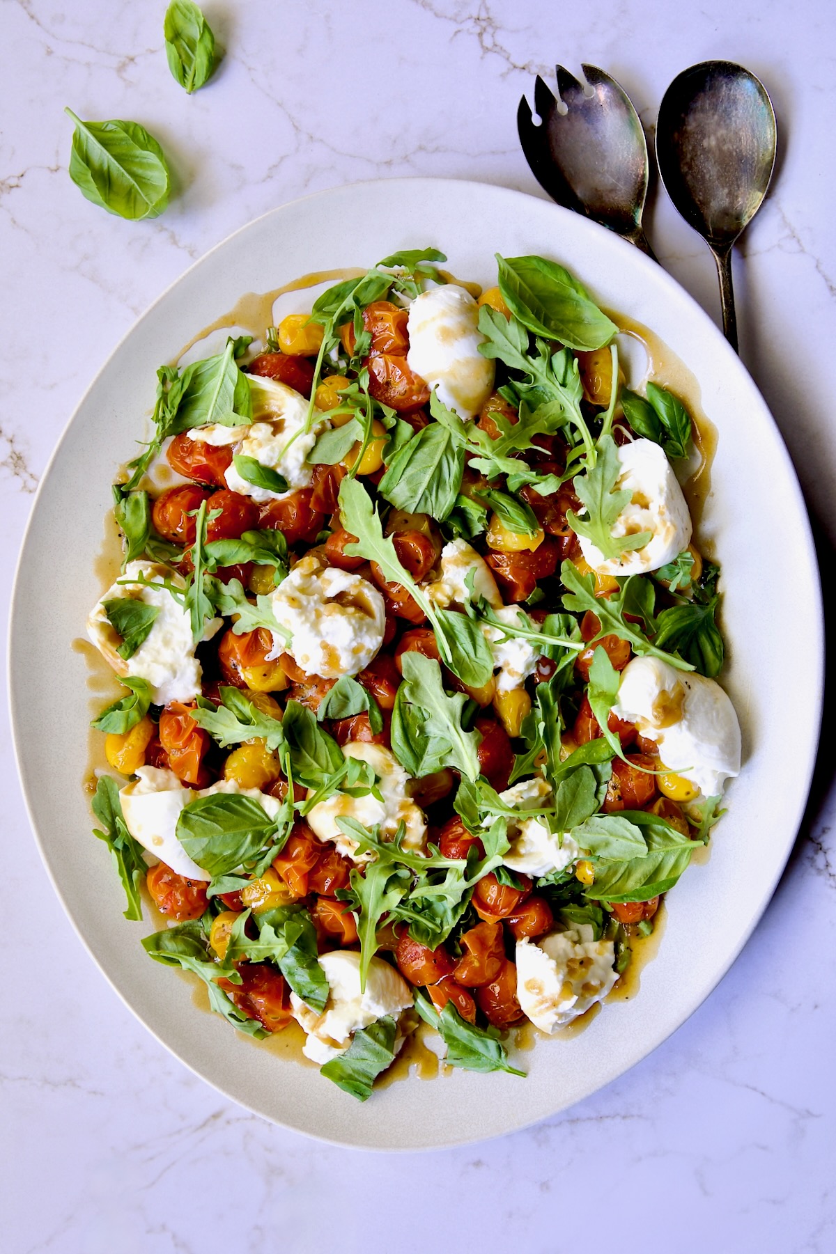 Roasted Cherry Tomato Burrata Caprese Salad on white oval platter with serving utensils.
