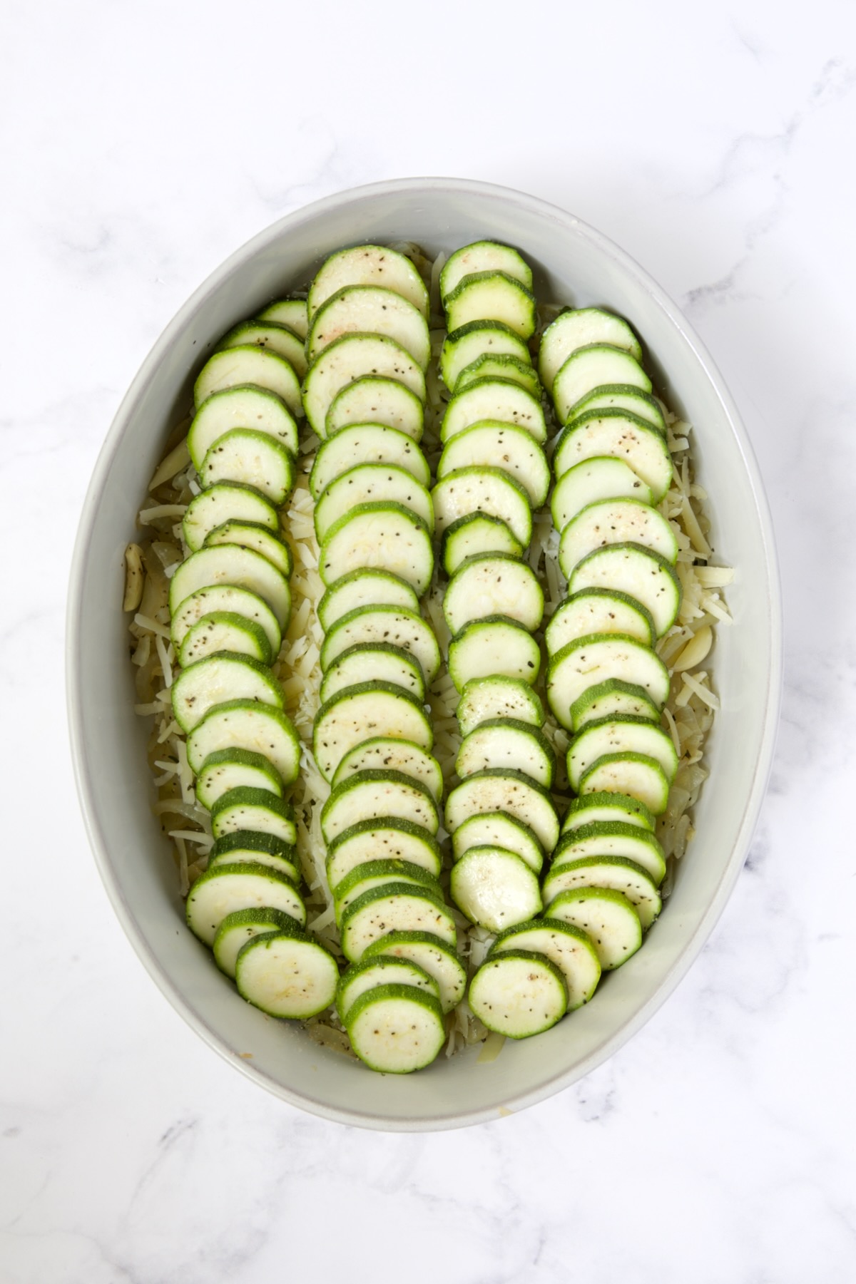 Sliced zucchini added to oval baking dish with the cheese and onion mixture.