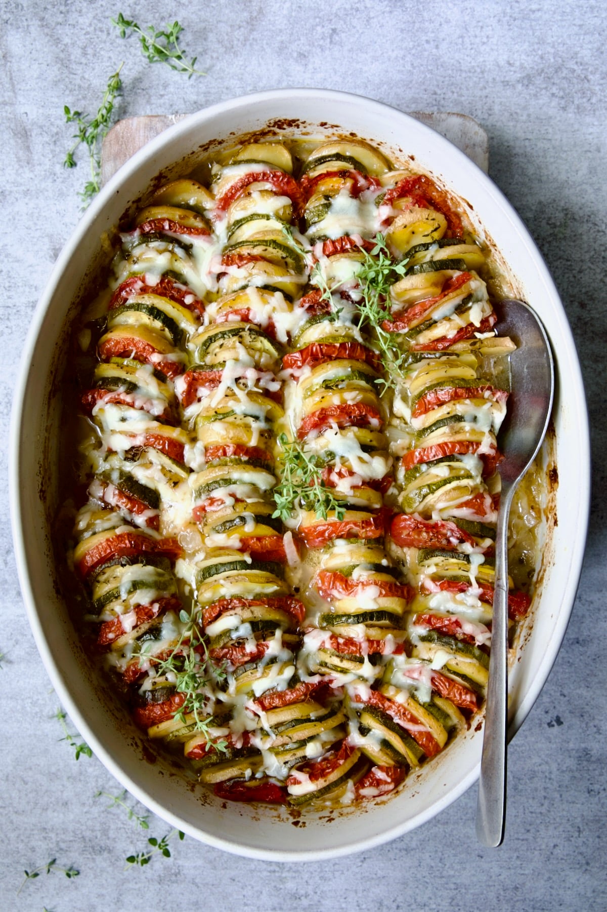 Vegetable Tian in oval baking dish with serving spoon garnished with fresh thyme.