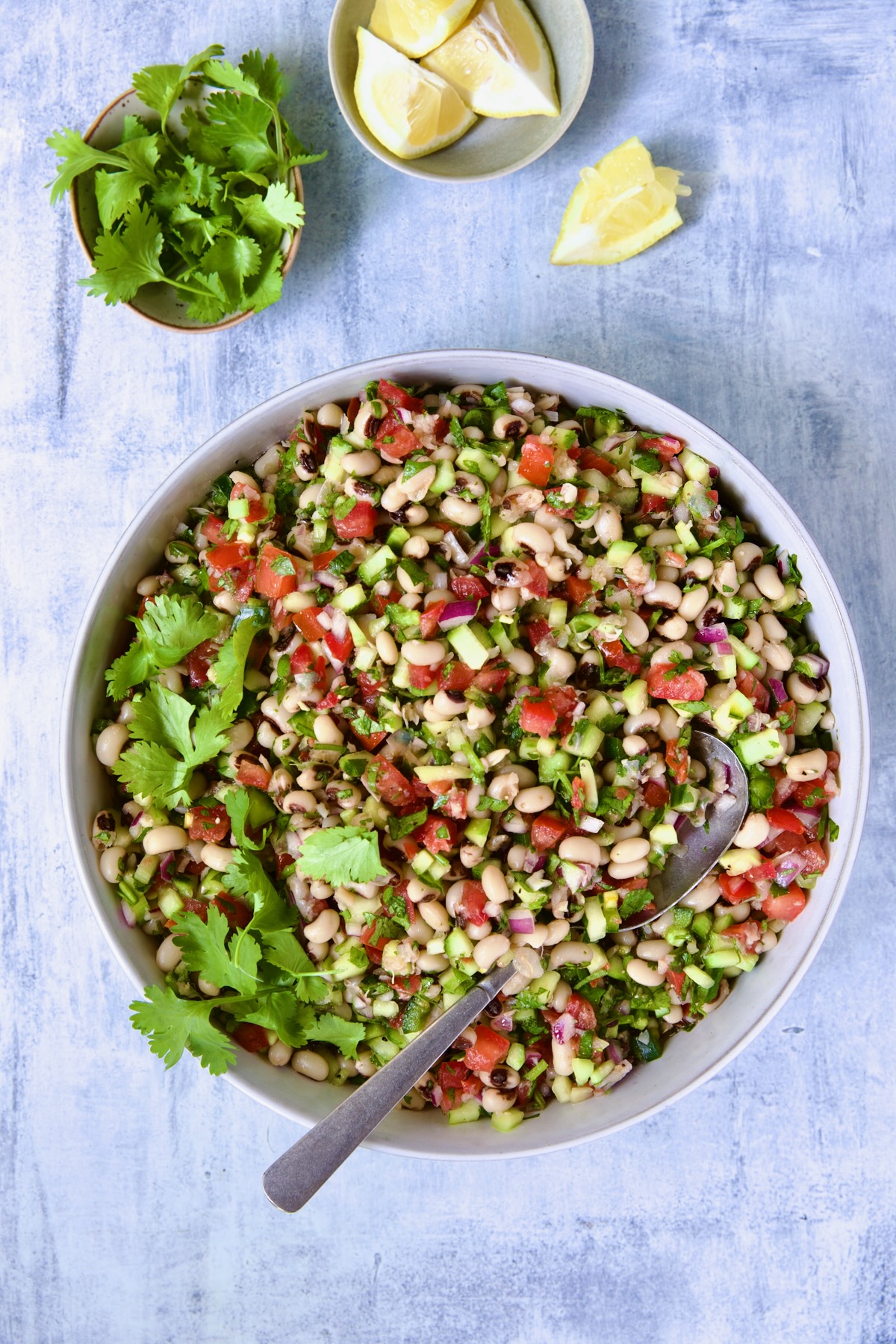 Black-Eyed Pea Salad in serving bowl with serving spoon and cilantro sprigs and lemon wedges in small bowls.