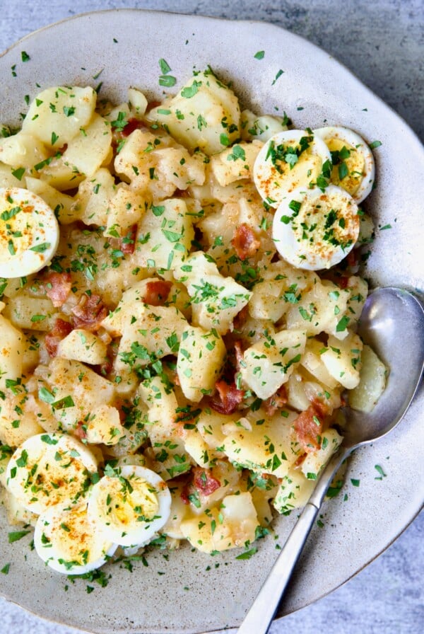 German Potato Salad in round earthenware bowl garnished with egg slices and fresh parsley with serving spoon.