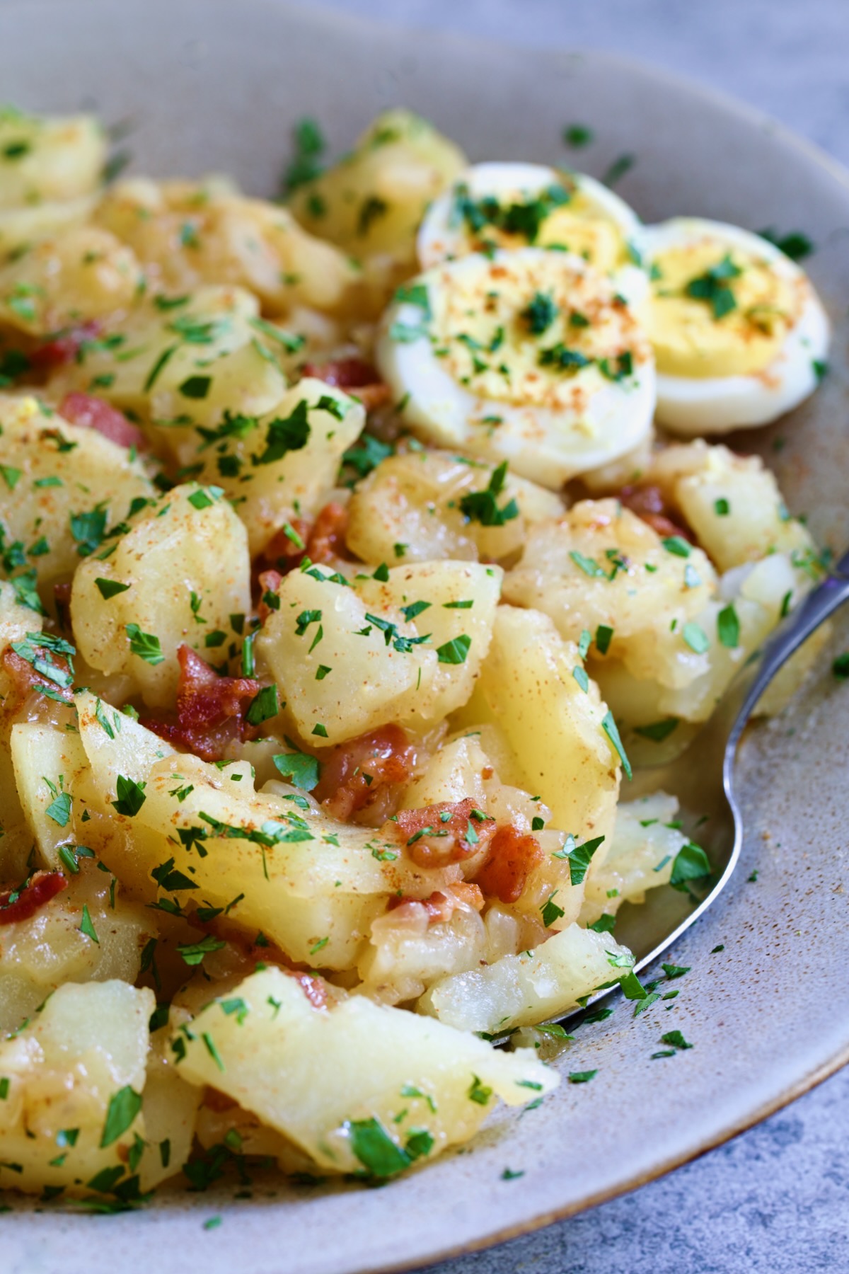 German Potato Salad in round earthenware bowl garnished with egg slices and fresh parsley with serving spoon.