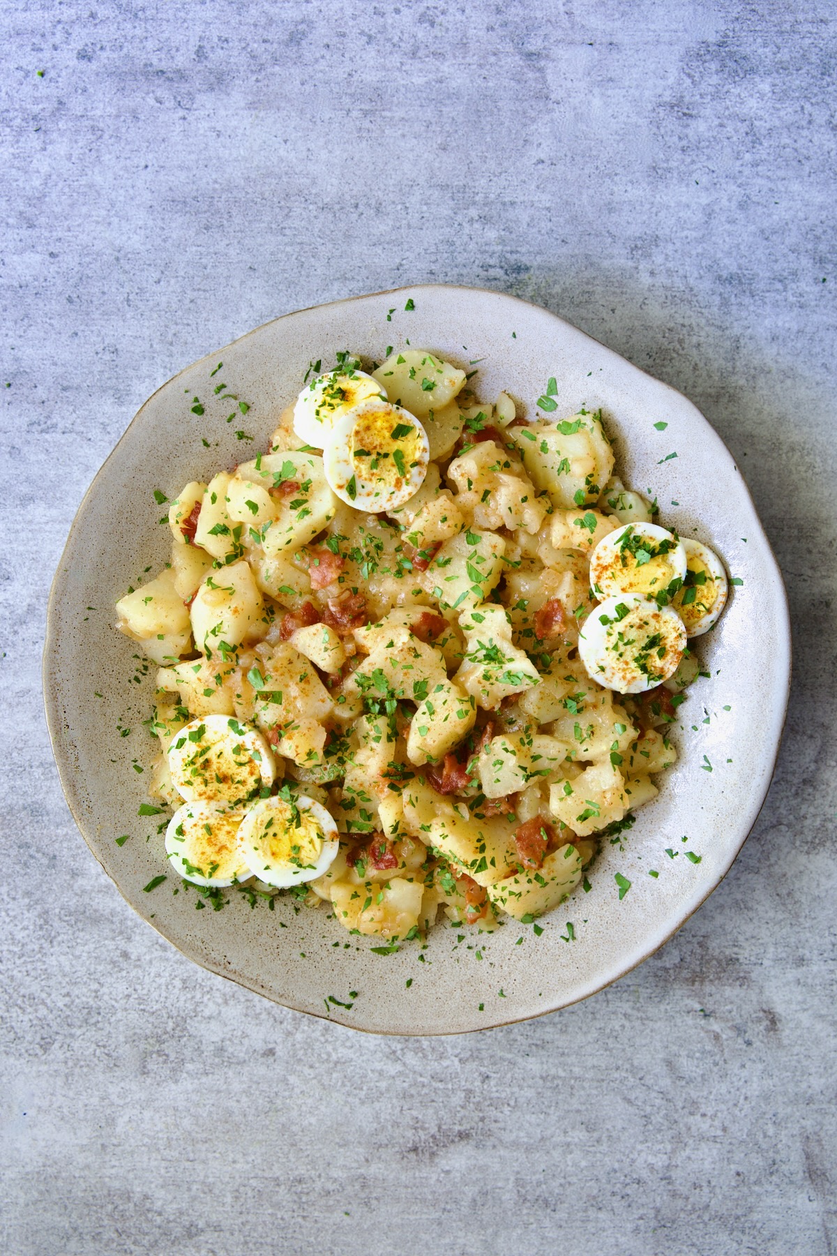 German Potato Salad in round earthenware bowl garnished with egg slices and fresh parsley.