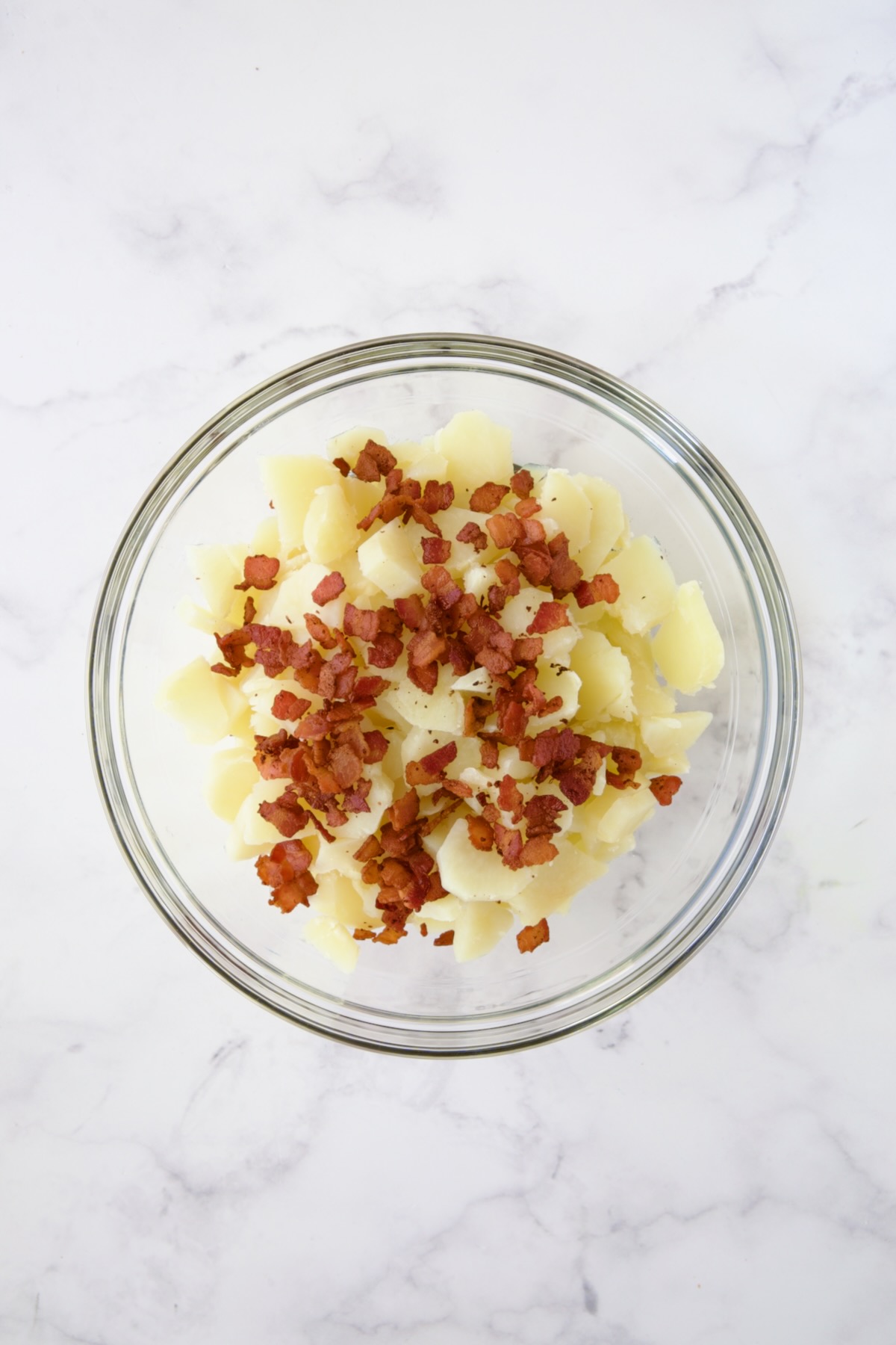 Cooked, sliced potatoes in glass bowl with bacon crumbled over the top.
