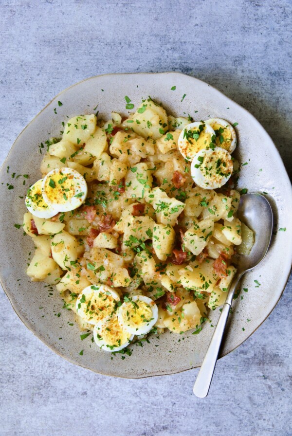 German Potato Salad in round earthenware bowl garnished with egg slices and fresh parsley with serving spoon.