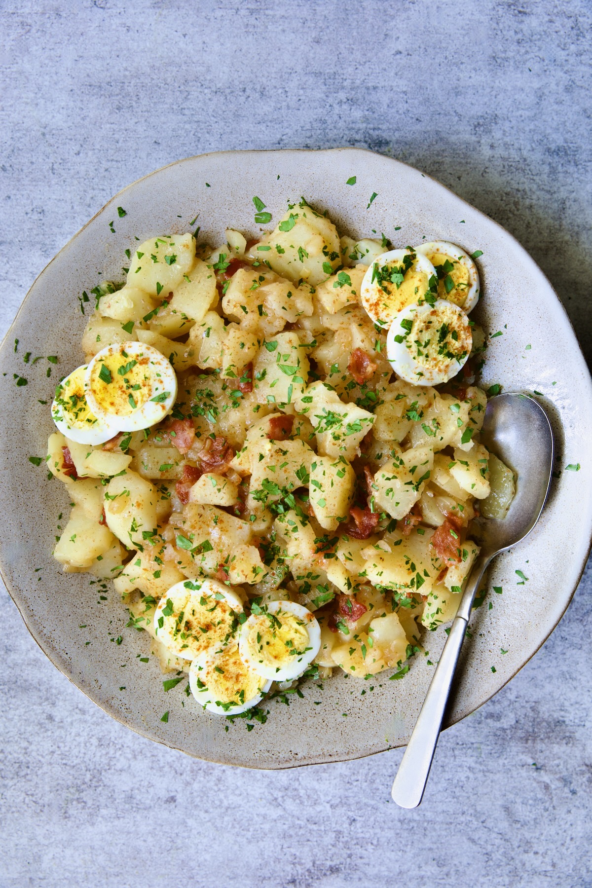 German Potato Salad in round earthenware bowl garnished with egg slices and fresh parsley with serving spoon.
