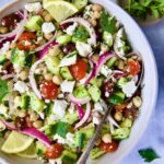 Greek Cucumber Salad in white bowl with serving spoon with oregano and parsley scattered about.