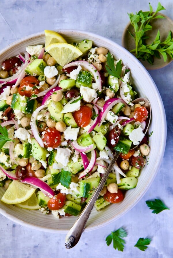 Greek Cucumber Salad in white bowl with serving spoon with oregano and parsley scattered about.