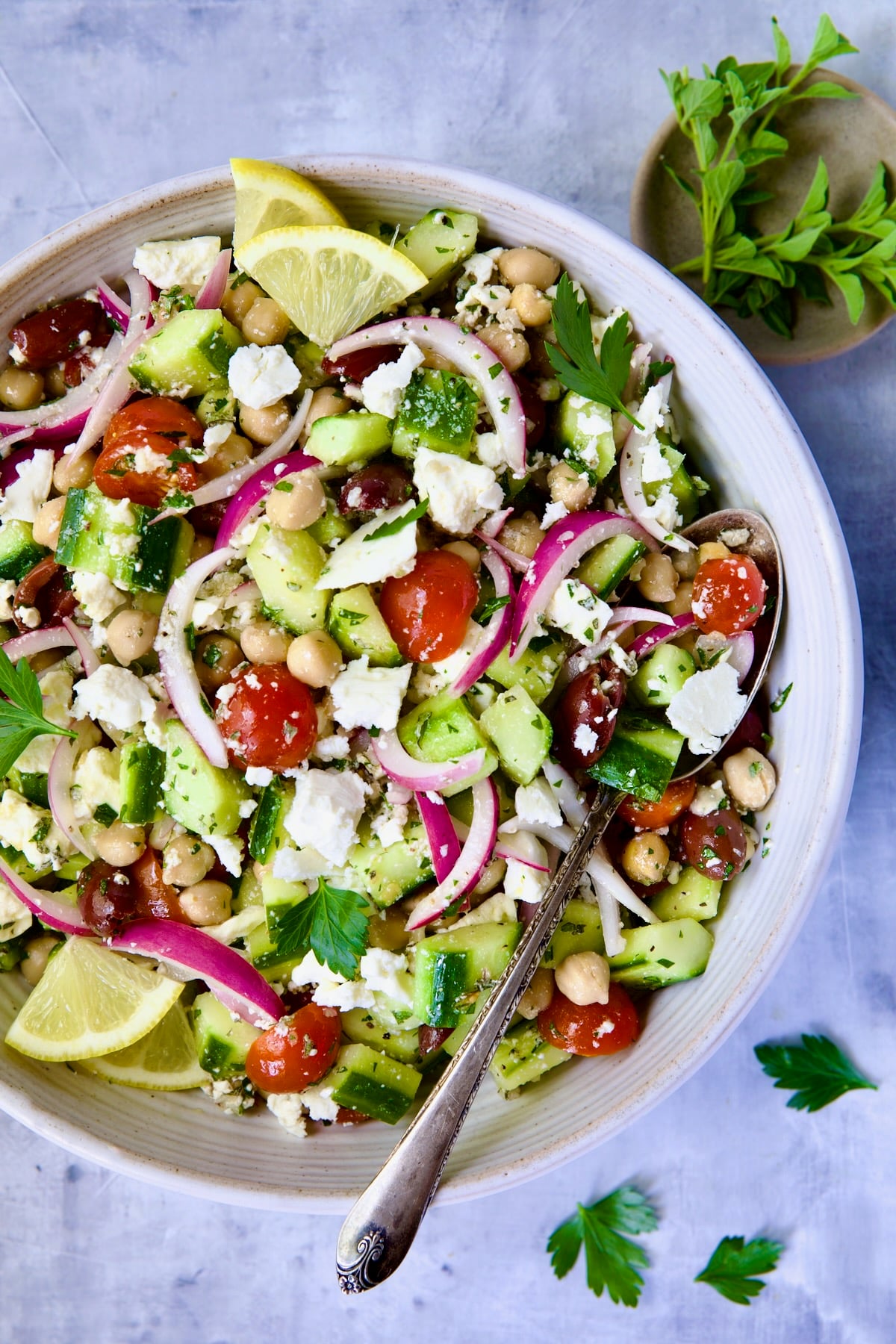 Greek Cucumber Salad in white bowl with serving spoon with oregano and parsley scattered about.