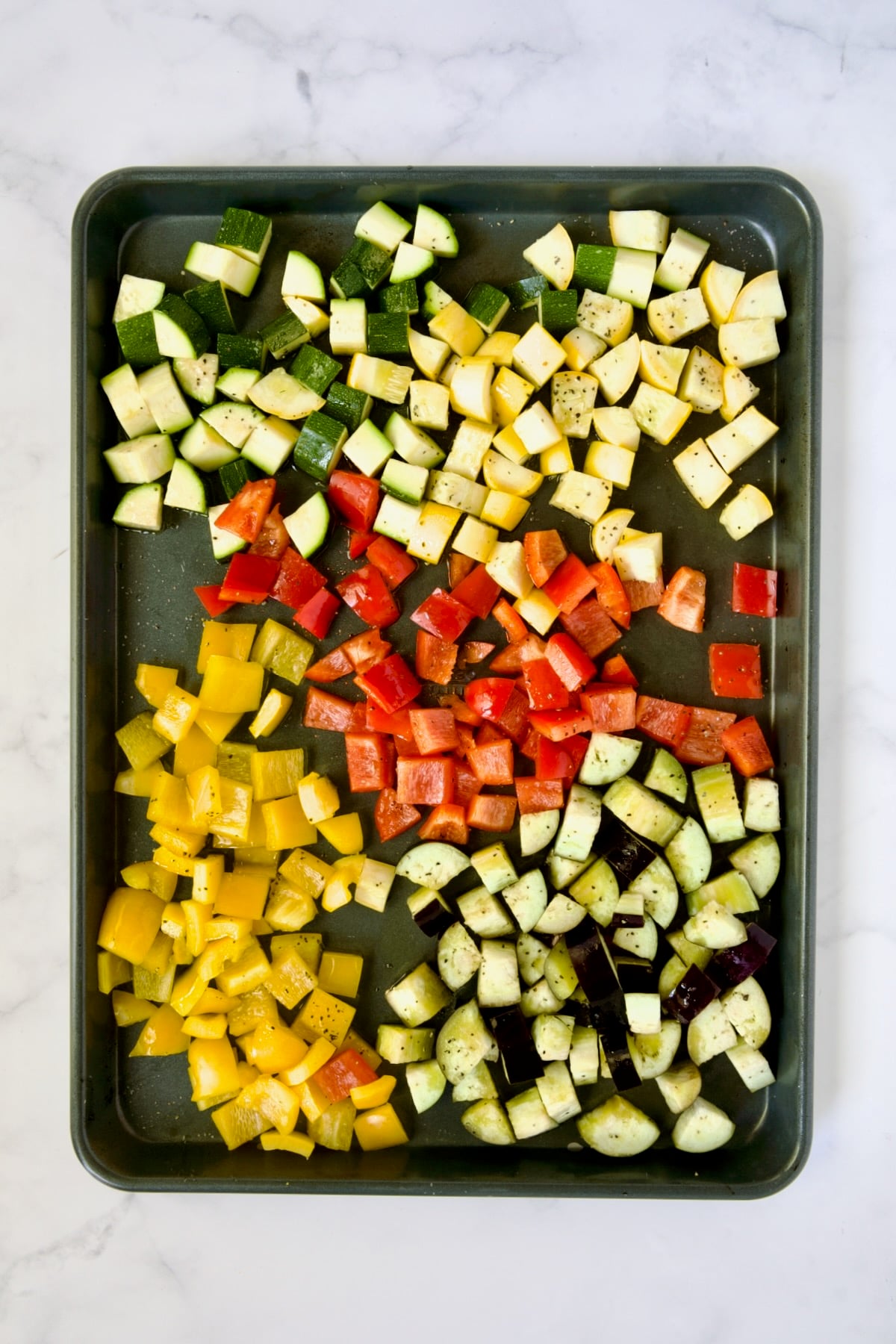 Cubed zucchini, yellow squash, eggplant, yellow and red bell pepper on rimmed sheet pan before being roasted.