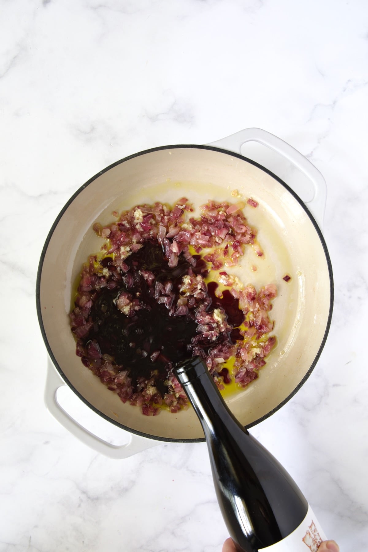 Red wine being poured onto onions from bottle into white enamel cast iron skillet.