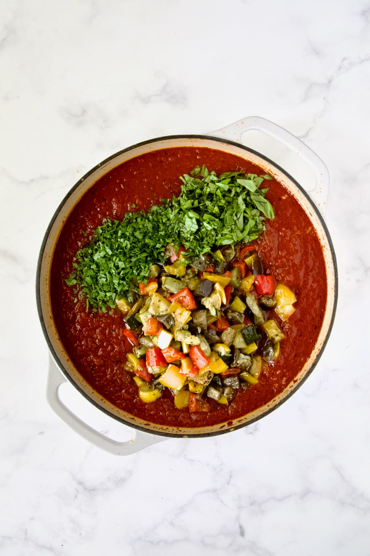 Roasted vegetables, parsley and fresh basil added to tomato sauce in white enamel cast iron skillet.