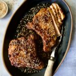 Two Asian Pork Chops on black oval platter garnished with sesame seeds with one pork chop partially sliced.