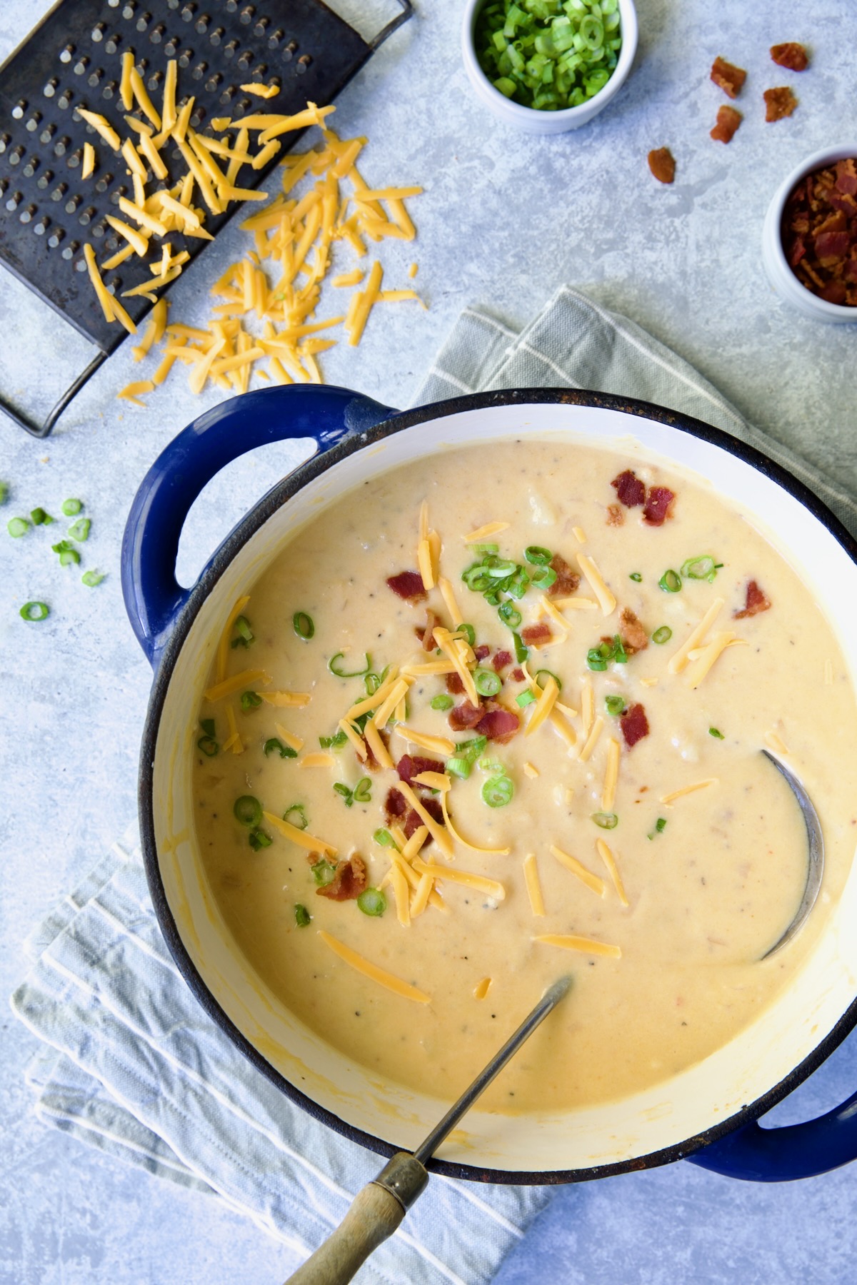 Cheddar Bacon Potato Soup in blue Dutch oven with soup ladle and cheese, scallion and bacon scattered about.