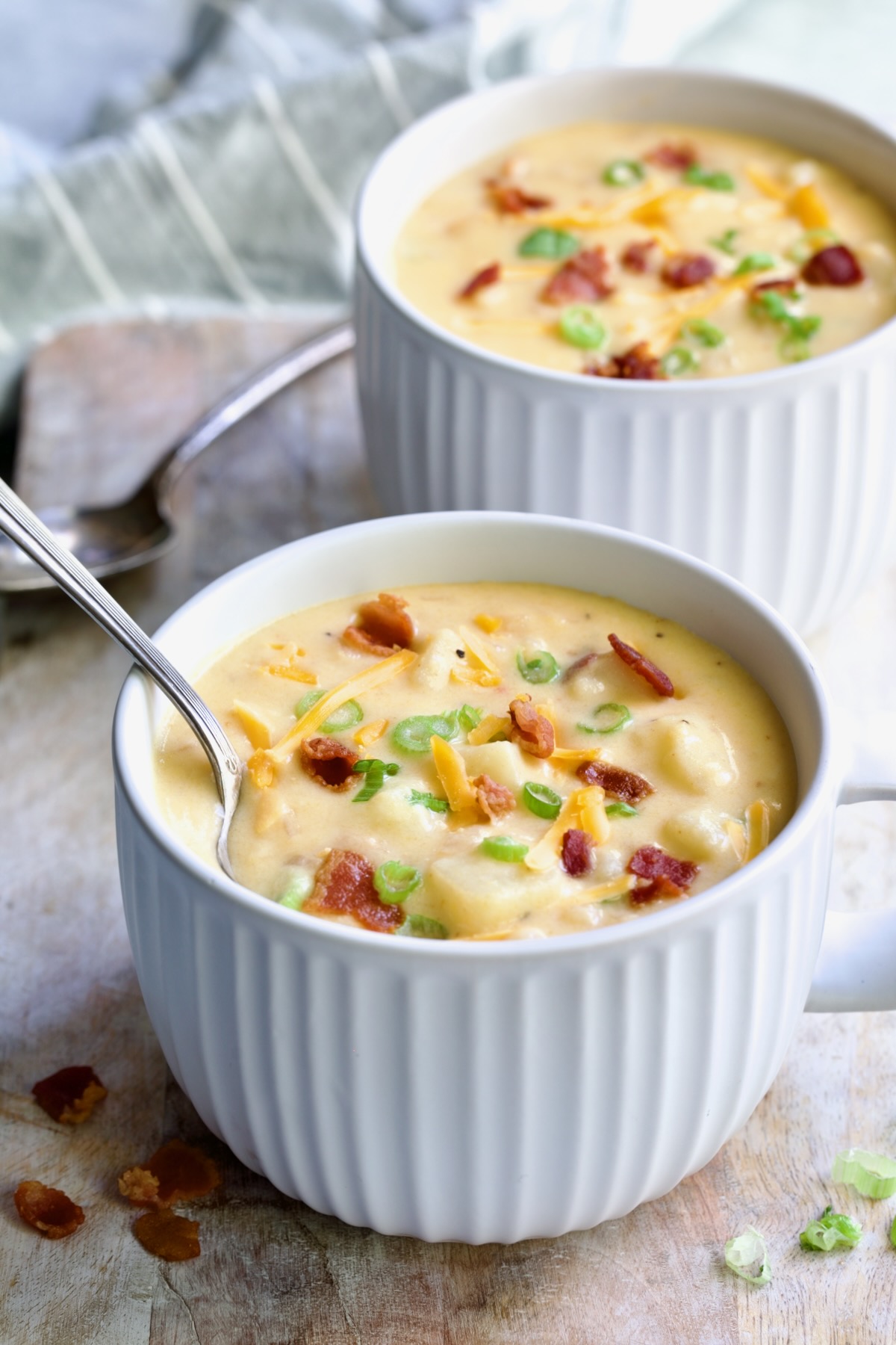Two large soup mugs of Cheddar Bacon Potato Soup garnished with bacon, scallion and Cheddar cheese.
