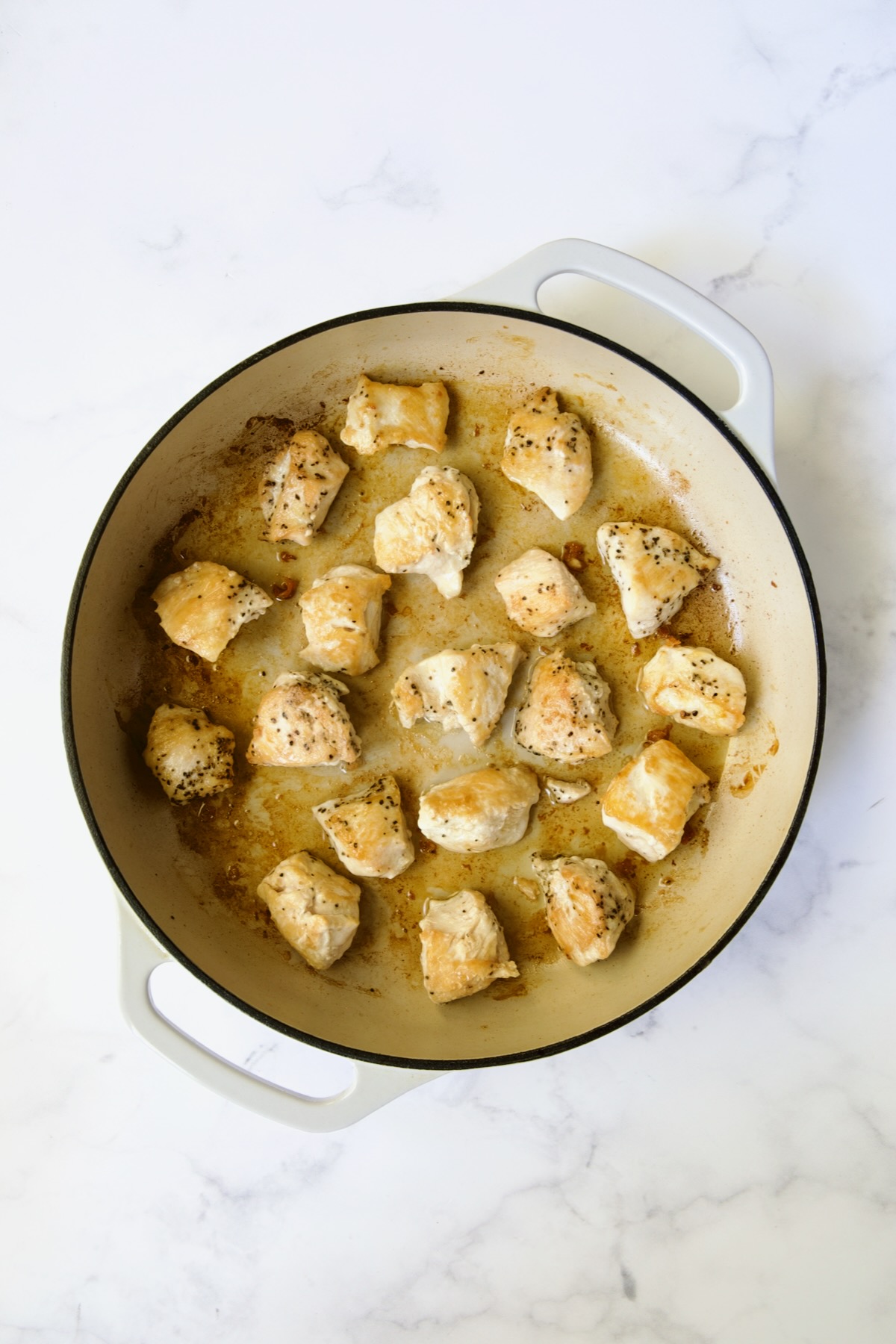 Browned cubed chicken in white enamel cast iron skillet.