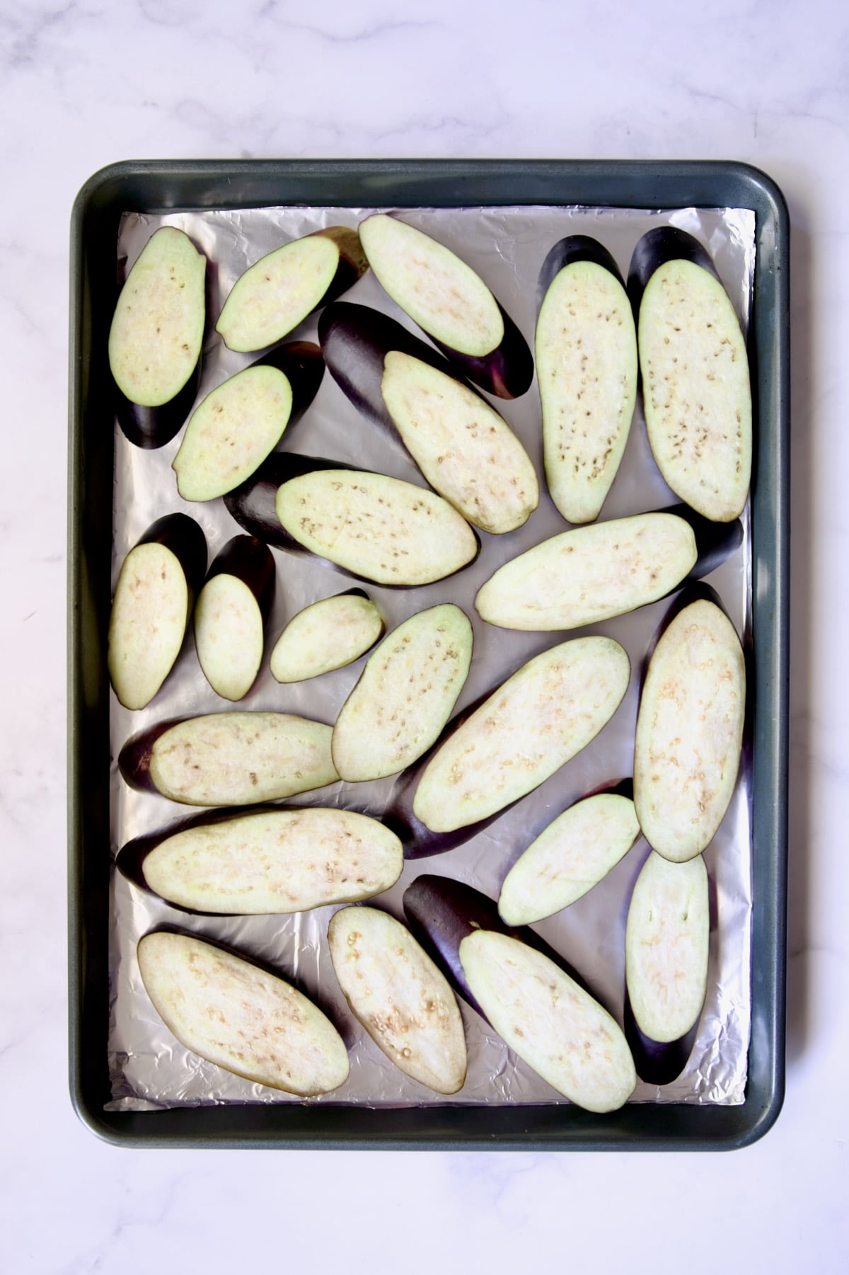 Slices of eggplant cut on the diagonal on aluminum foil-lined rimmed sheet pan.