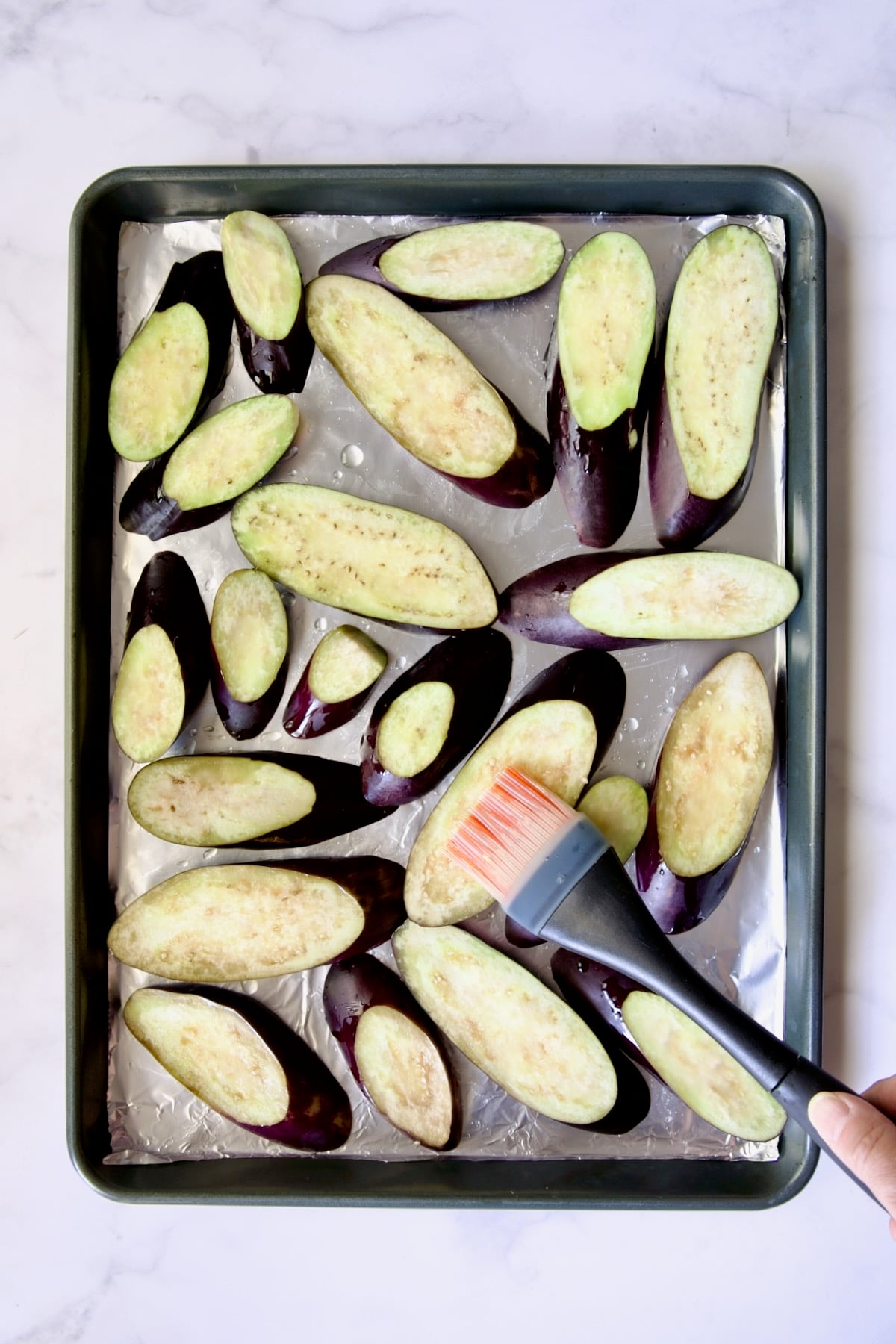 Slices of eggplant cut on the diagonal on aluminum foil-lined rimmed sheet pan being brushed with canola oil.