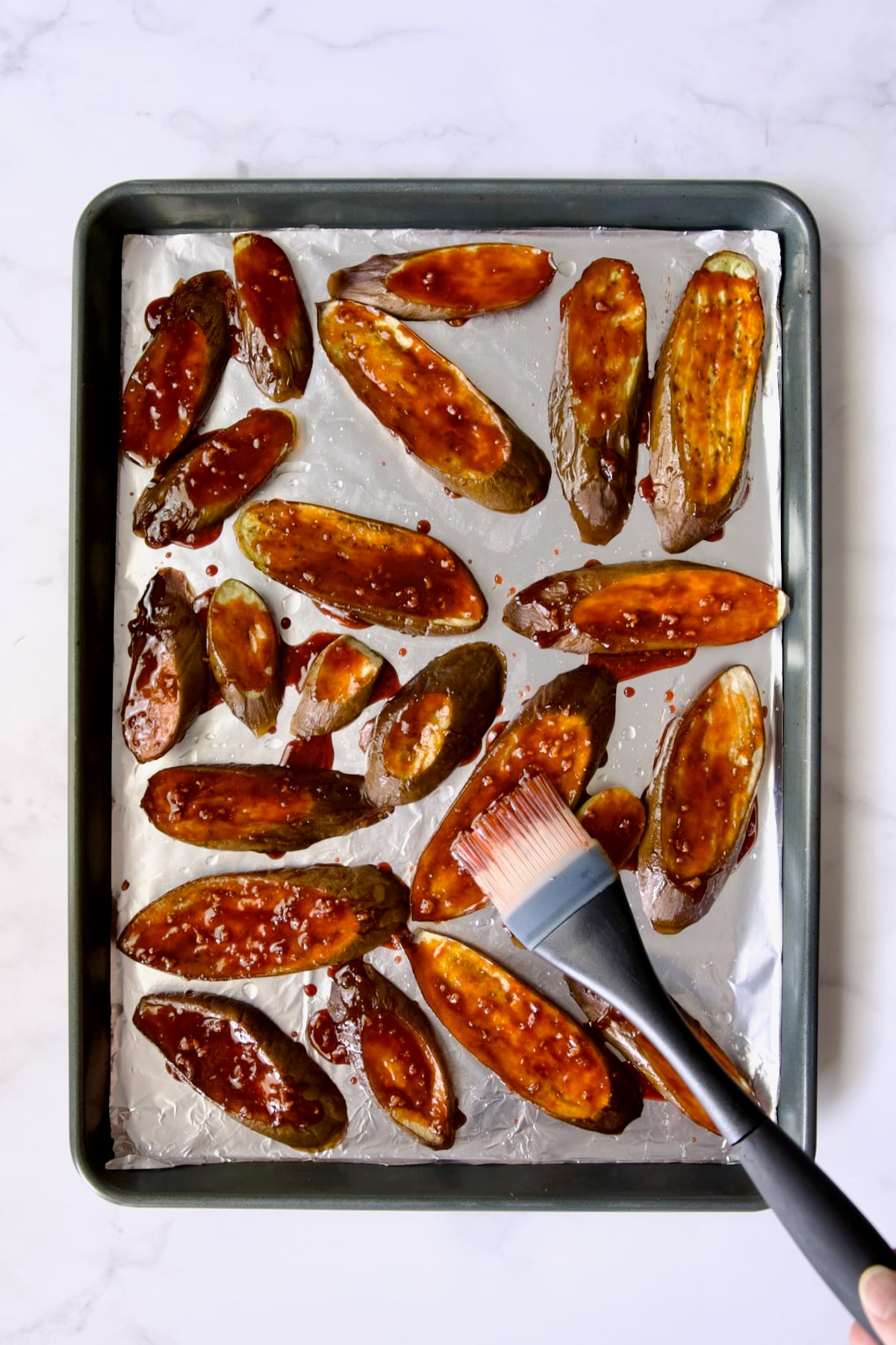 Slices of eggplant cut on the diagonal on aluminum foil-lined rimmed sheet pan being brushed with sauce.
