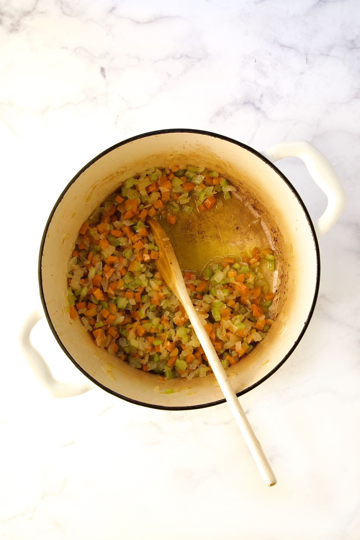 Cooked onions, carrots and celery in white Dutch oven with wooden spoon.