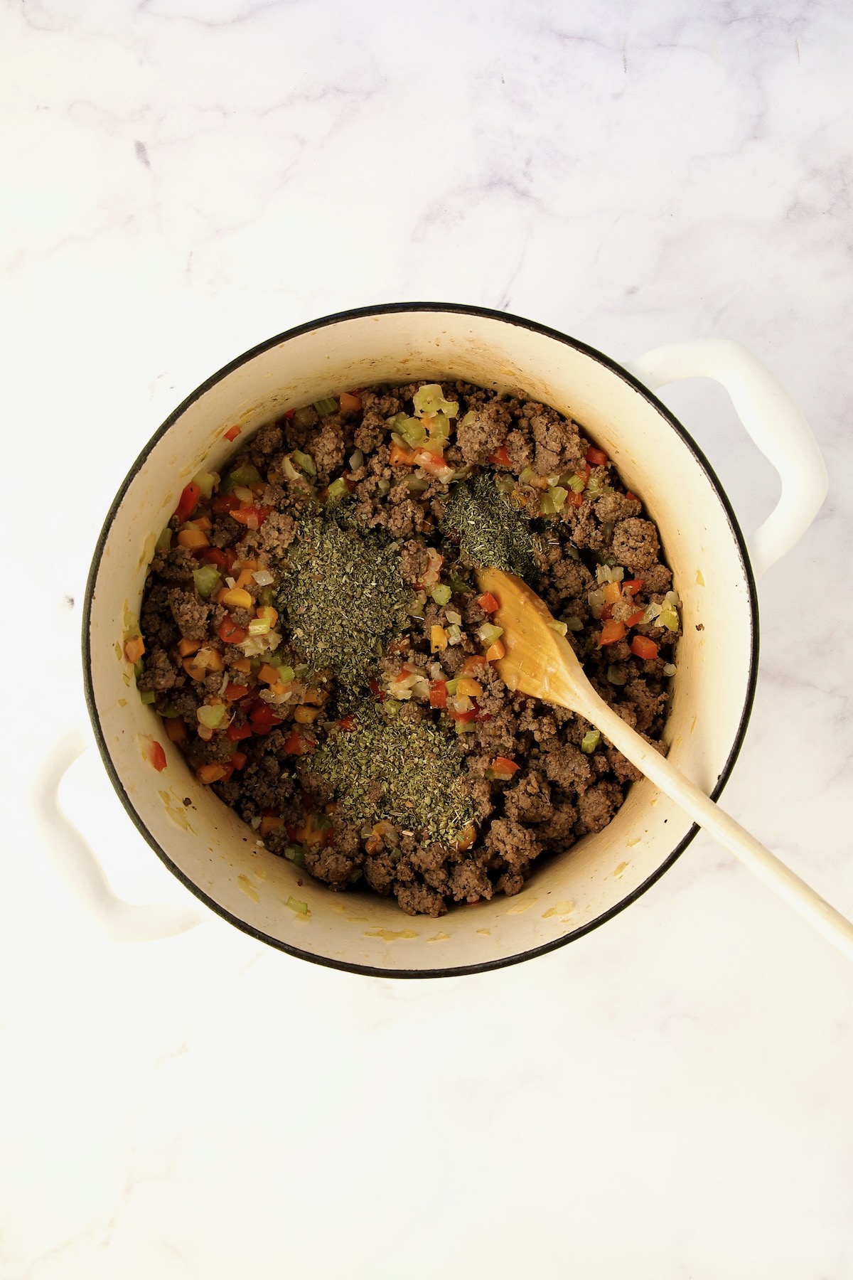 Dried herbs added to cooked ground beef, onions, carrots and celery in white Dutch oven.