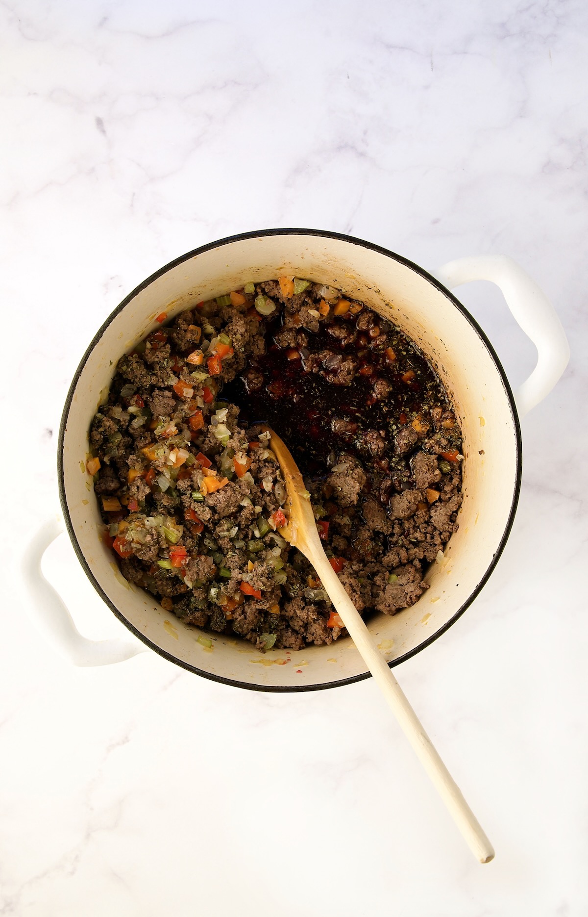 Red wine added to cooked ground beef, onions, carrots and celery in white Dutch oven being stirred with wooden spoon.